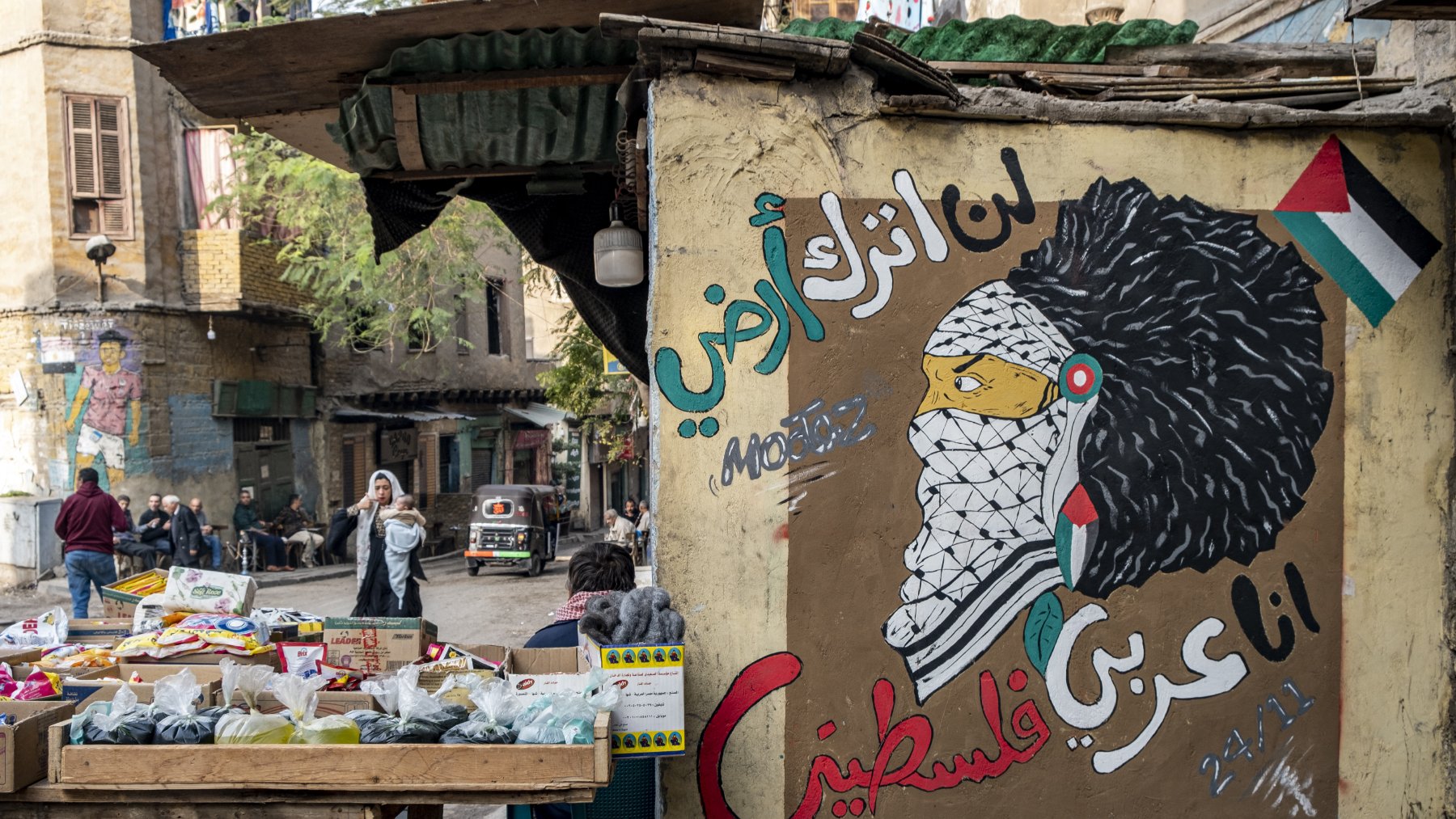 L'image montre un mur peint avec une fresque représentant une figure symbolique, probablement inspirée par la culture palestinienne. Le personnage porte un keffieh et a une chevelure élaborée. À côté, il y a des inscriptions en arabe qui expriment des sentiments de résistance et d'identité palestinienne. En avant-plan, il y a un marché avec des étals de fruits et légumes, et une rue animée où l'on peut apercevoir des passants et des véhicules. L'ambiance reflète un mélange de culture, d'art et de vie urbaine.