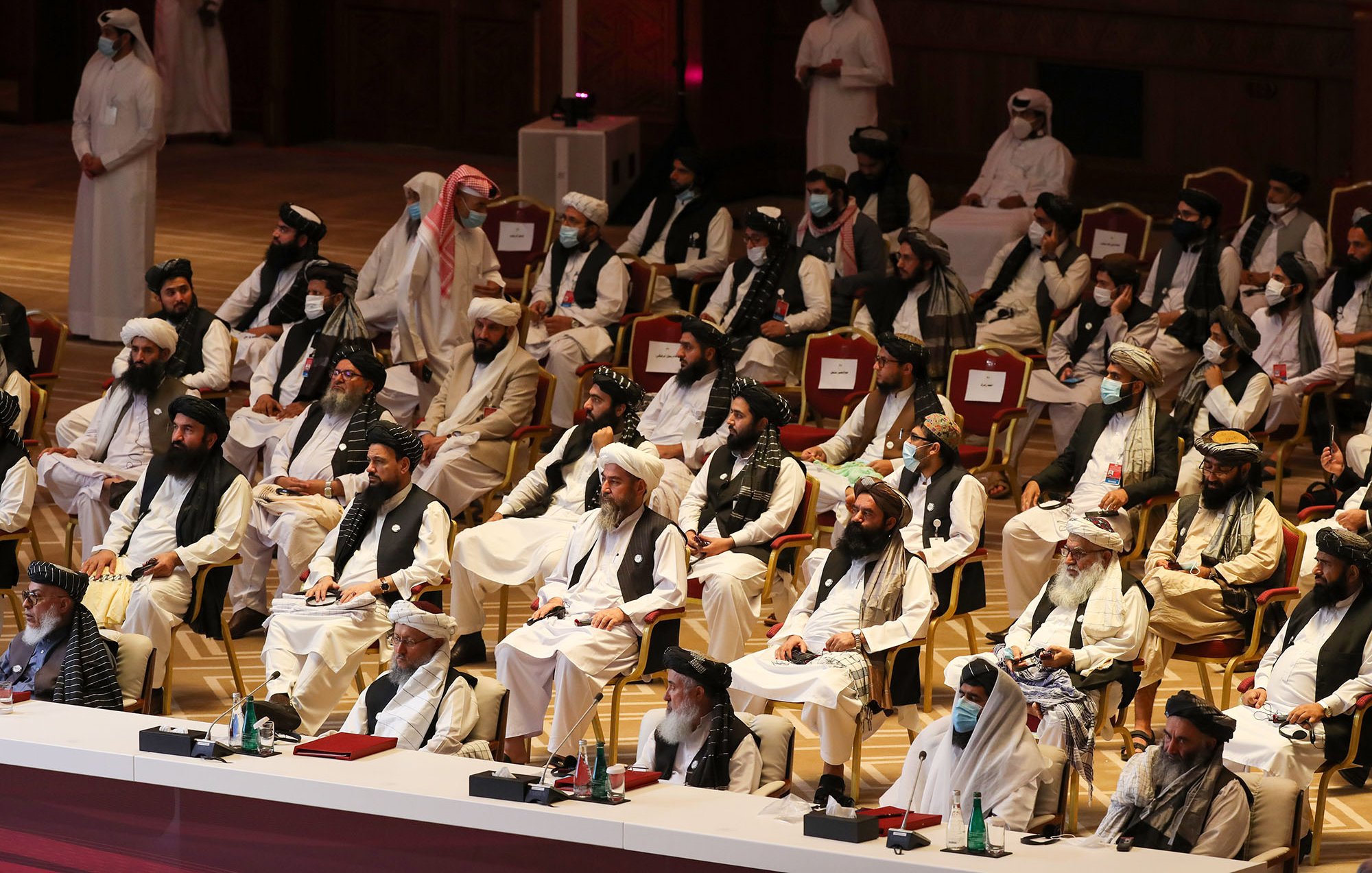 L'image montre un grand rassemblement de personnes assises dans une salle. Les participants sont principalement vêtus de vêtements traditionnels, tels que des turbans et des robes longues. Certains portent des masques. Les gens semblent attentifs et concentrés sur l'événement en cours. L'environnement est formel, avec des sièges disposés en rangées, et une scène à l'avant. L'atmosphère générale évoque une réunion officielle ou un congrès.