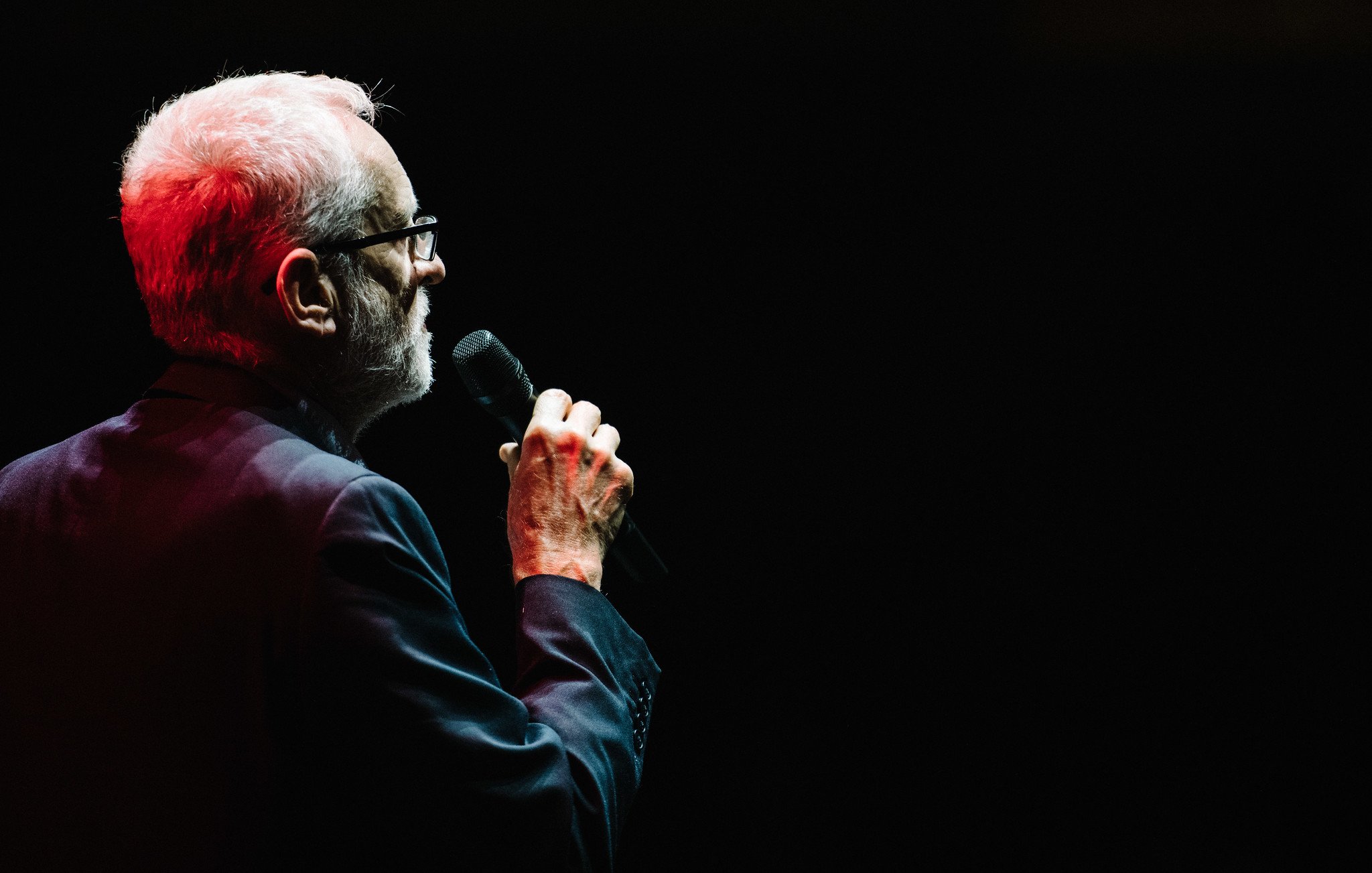 L'image montre un homme âgé, vu de profil, debout sur scène. Il tient un micro à la main et regarde vers le public. Sa chevelure est grise et il porte des lunettes. Il est habillé d'un costume sombre, ce qui contraste avec l'éclairage rouge qui accentue son visage et ses mains. L'arrière-plan est sombre, ce qui met en valeur sa silhouette et son expression. L'atmosphère semble formelle et engageante, suggérant qu'il est en train de donner un discours ou de chanter.