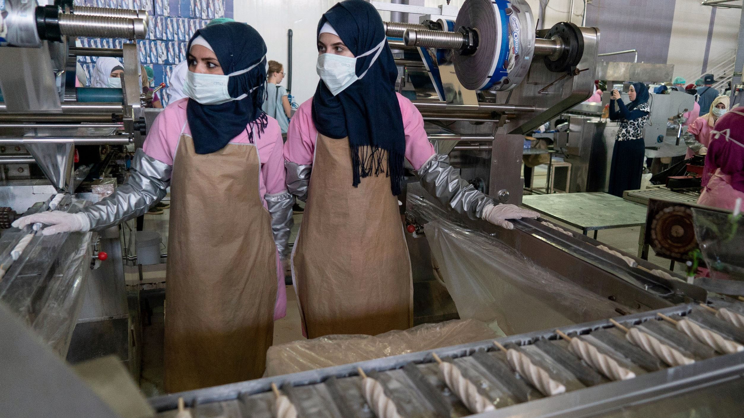 L'image montre deux femmes travaillant dans un environnement industriel. Elles portent des hijabs et des masques de protection. Elles sont habillées de vêtements de travail, avec des blouses et des tabliers. À l'arrière-plan, on peut voir des machines et d'autres personnes également en train de travailler, ce qui indique qu'il s'agit probablement d'une usine ou d'un atelier de production. L'atmosphère semble axée sur le travail et la production.