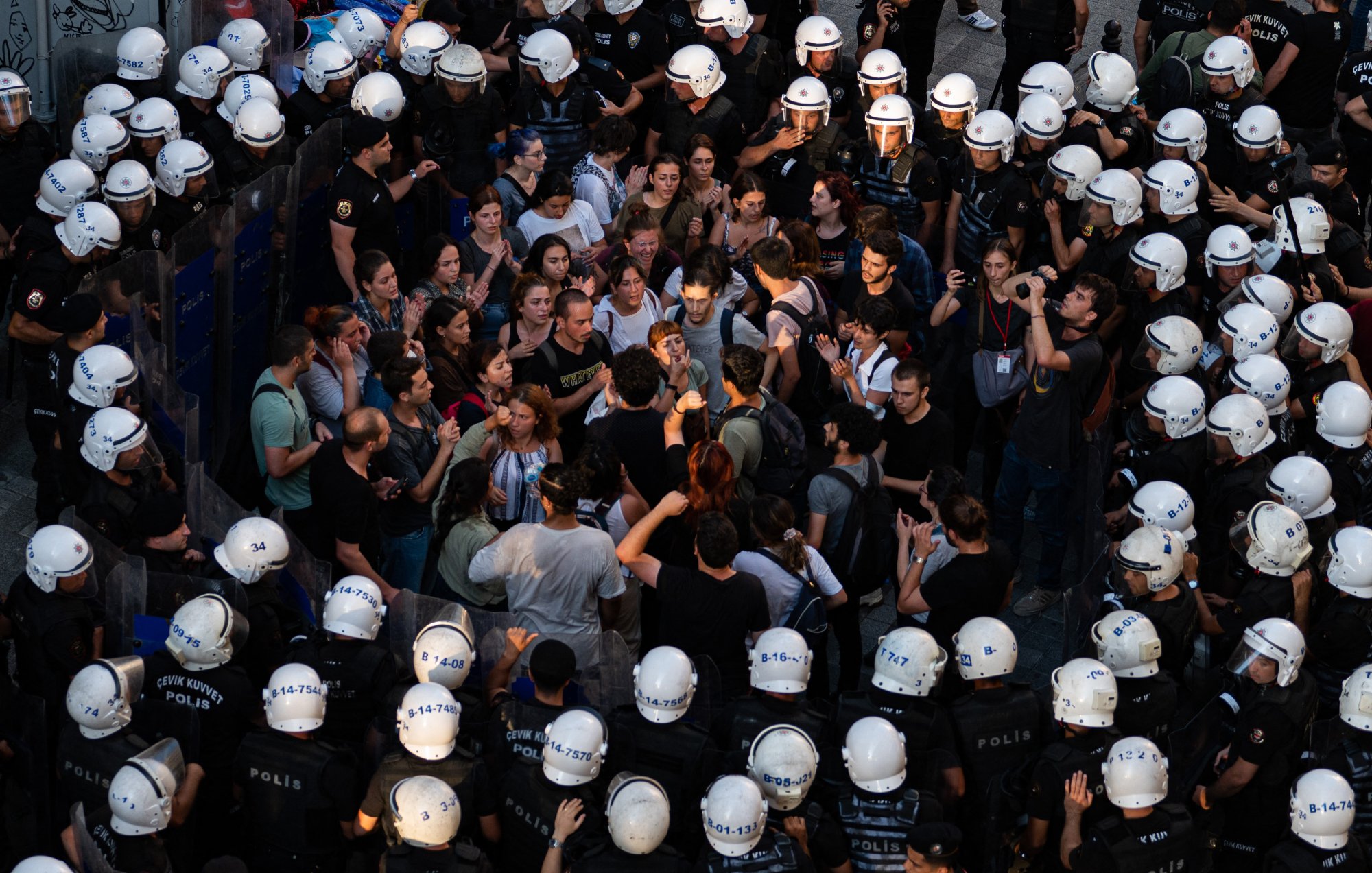 L'image montre une scène de manifestation où une foule de personnes se trouve face à des policiers en tenue anti-émeute. Les manifestants semblent discuter ou protester, tandis que les policiers forment une ligne serrée. L'atmosphère est tendue, et une partie de la foule semble concentrée sur un point, indiquant une interaction ou un affrontement verbal. Les casques des policiers et leur équipement renforcent l'impression de confrontation.