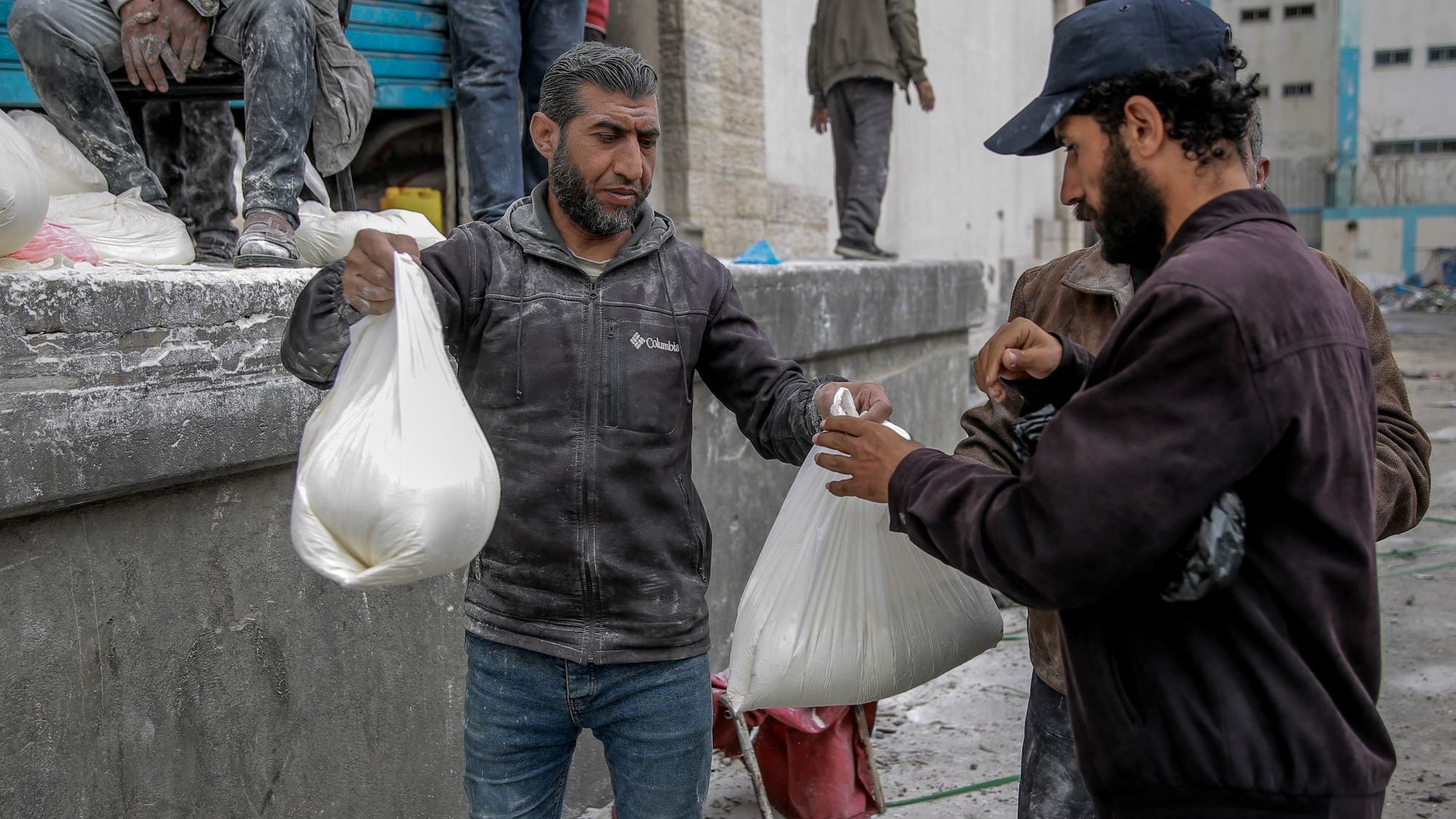 L'image montre des hommes dans un environnement urbain, apparemment en train de distribuer ou de recevoir des sacs. Ils semblent porter des vêtements de travail, certains étant couverts de poussière ou de matériau. L'ambiance suggère une activité liée à l'aide humanitaire ou à une distribution de biens, dans un contexte peut-être de reconstruction ou d'assistance. Les interactions entre les hommes semblent concentrées et sérieuses.