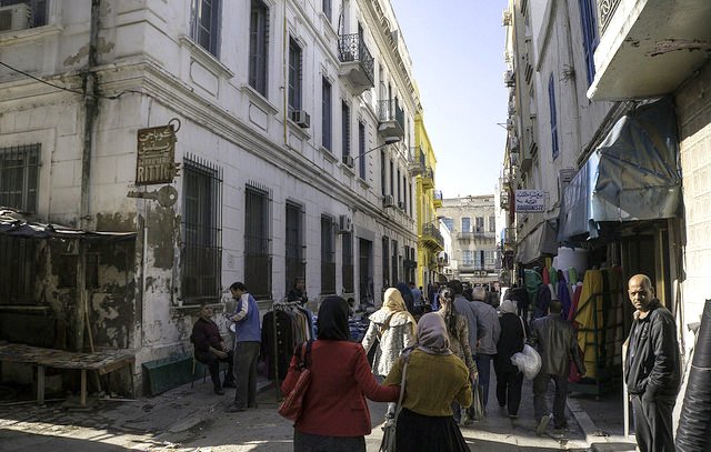 L'image montre une ruelle animée avec des bâtiments à l'architecture typique, dont certains sont peints en blanc et en jaune. On peut voir des gens marchant dans la rue, dont des femmes portant des habits traditionnels. À gauche, il y a un marché ou des stands avec des marchandises. L'ambiance semble vivante et typique d'un quartier urbain. La lumière du soleil éclaire la scène, ajoutant une chaleur à l'atmosphère.