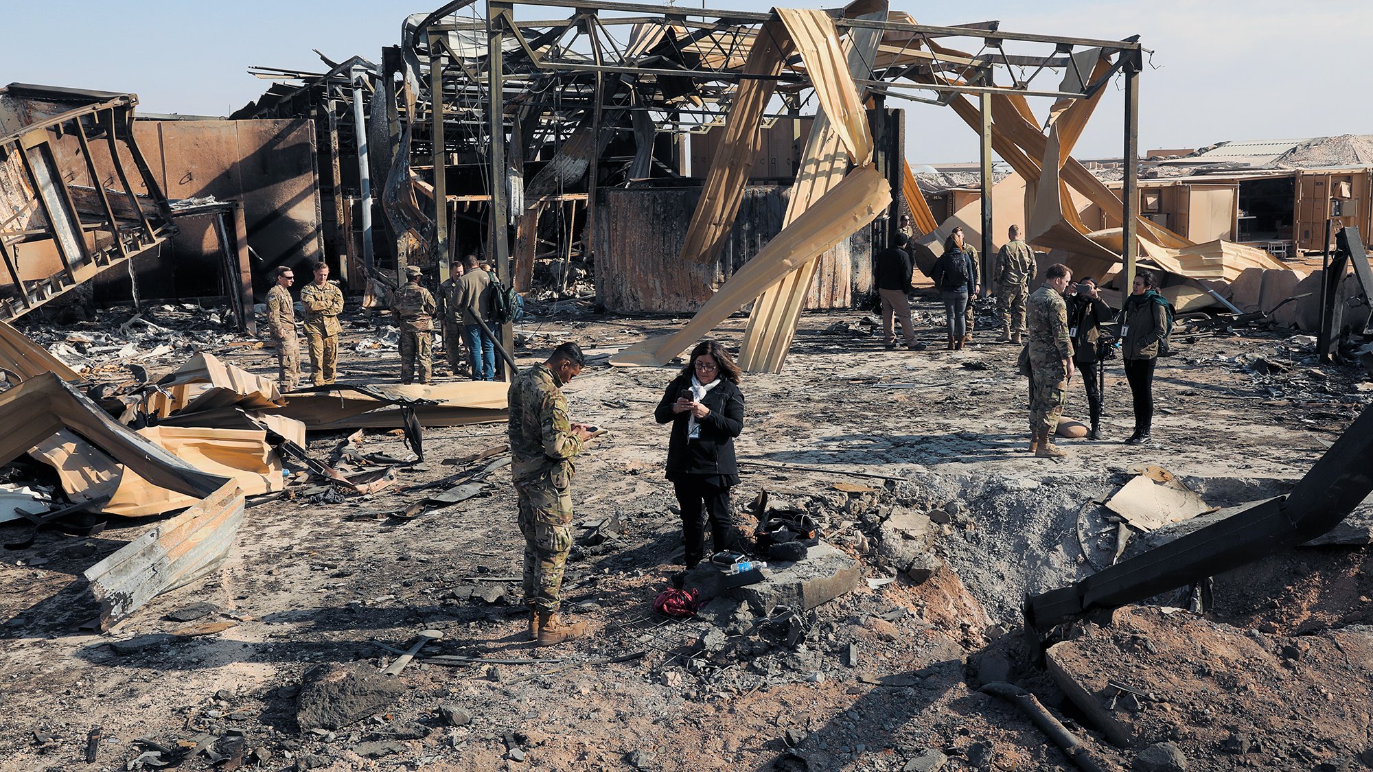 L'image montre une scène de destruction, probablement après un bombardement ou un incendie. On peut voir des débris et des structures en ruines, avec des panneaux de bois fissurés. Plusieurs militaires et civils se trouvent sur le site, certains en train de prendre des notes ou d'observer les dégâts. L'atmosphère semble grave, soulignant les conséquences d'un conflit. Le ciel est clair, contrastant avec le désastre au sol.