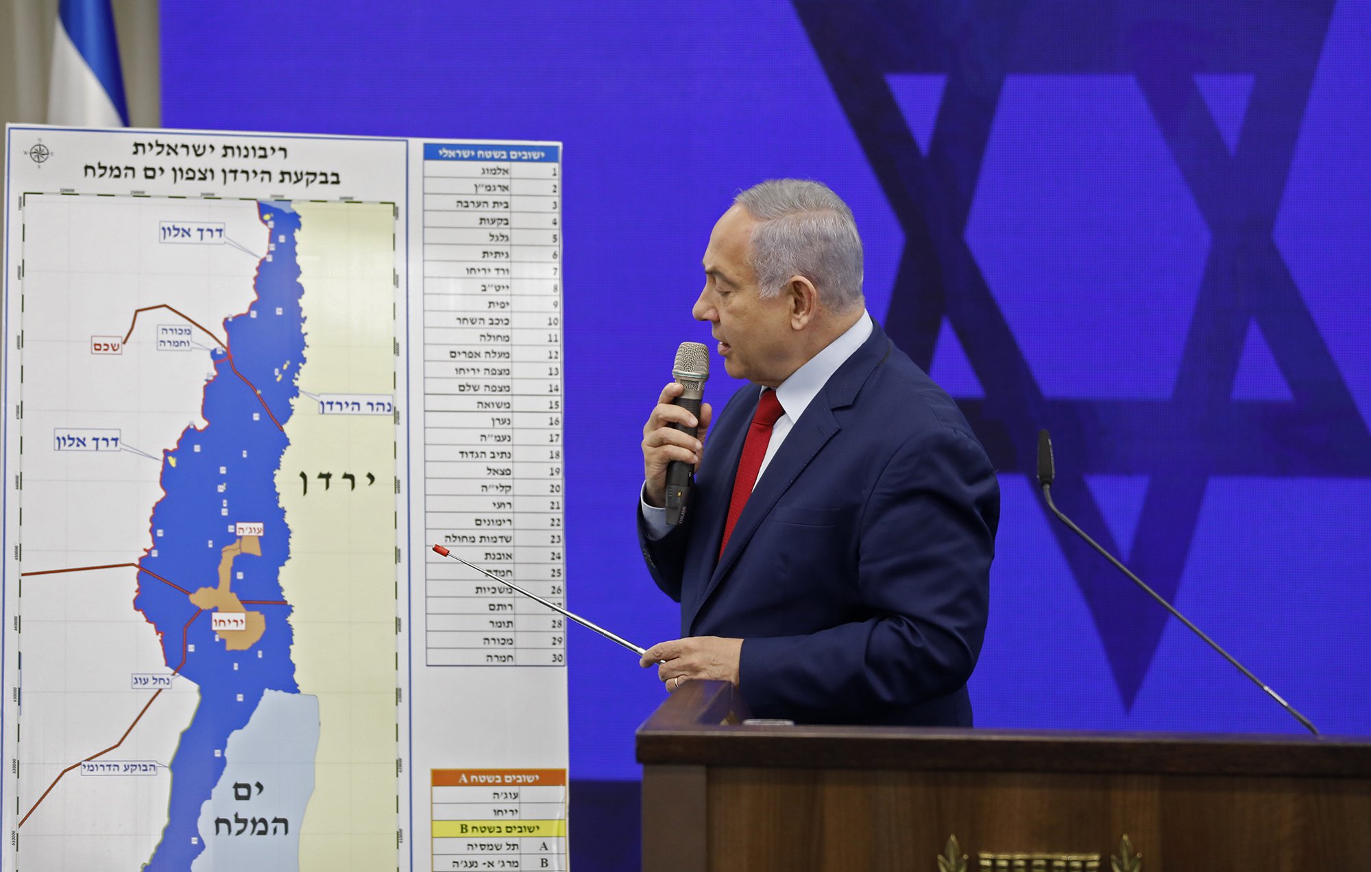 L'image montre un homme debout devant un tableau graphique. Il semble s'adresser à un auditoire, tenant un pointeur dans sa main. Le tableau derrière lui illustre une carte, probablement liée à une question de territoire, avec des annotations en hébreu. On aperçoit également un symbole du judaïsme en arrière-plan. L'homme porte une cravate rouge et un costume sombre.