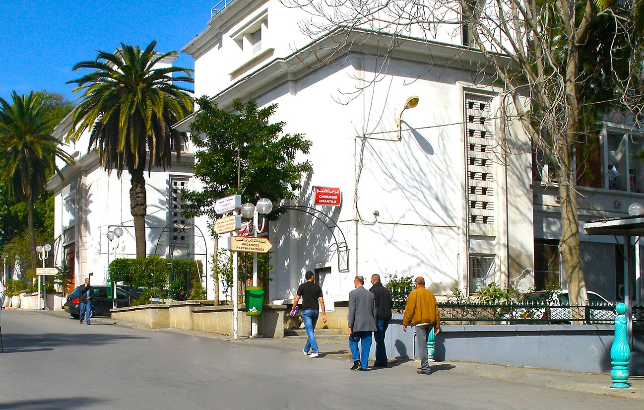 L'image montre une rue dégagée avec des bâtiments blancs et modernes de chaque côté. Des palmiers bordent le chemin, ajoutant une touche tropicale à l'environnement. On peut voir plusieurs personnes marchant le long de la rue, certaines en tenue traditionnelle. Le ciel est bleu et ensoleillé, ce qui donne une ambiance agréable et décontractée à la scène. Les façades des bâtiments présentent une architecture typique de la région, avec des détails subtils comme des lampadaires et des panneaux indiquant des directions.