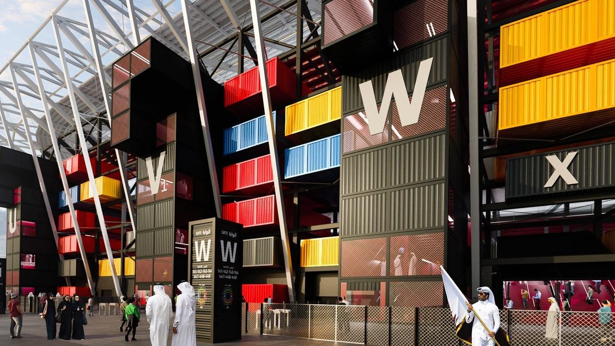 L'image montre une vue d'un bâtiment moderne, probablement un stade ou un centre événementiel, avec une architecture unique. Les murs extérieurs sont composés de containers colorés, alternant des teintes de jaune, rouge et bleu, donnant un aspect dynamique et contemporain. On distingue également des personnes en vêtements traditionnels se tenant près de l'entrée, ainsi que des éléments de sécurité. Le ciel est légèrement nuageux, ajoutant une ambiance particulière à la scène.