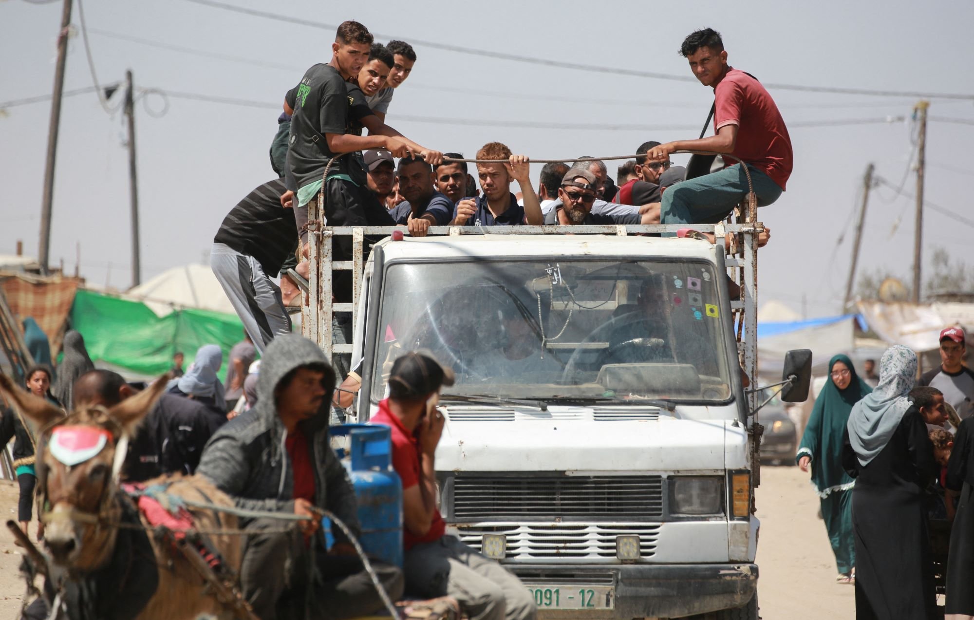 L'image montre un groupe de personnes assises sur le toit d'un camion, tandis que d'autres se trouvent à proximité, y compris des personnes à pied et une charrette tirée par un cheval. L'environnement semble être un endroit animé, avec des tentes et des structures visibles en arrière-plan. On peut voir des fils électriques et des montagnes de déchets, ce qui suggère une atmosphère informelle ou de camp. La scène dégage une impression de communauté et de vie quotidienne dans un contexte peut-être difficile.