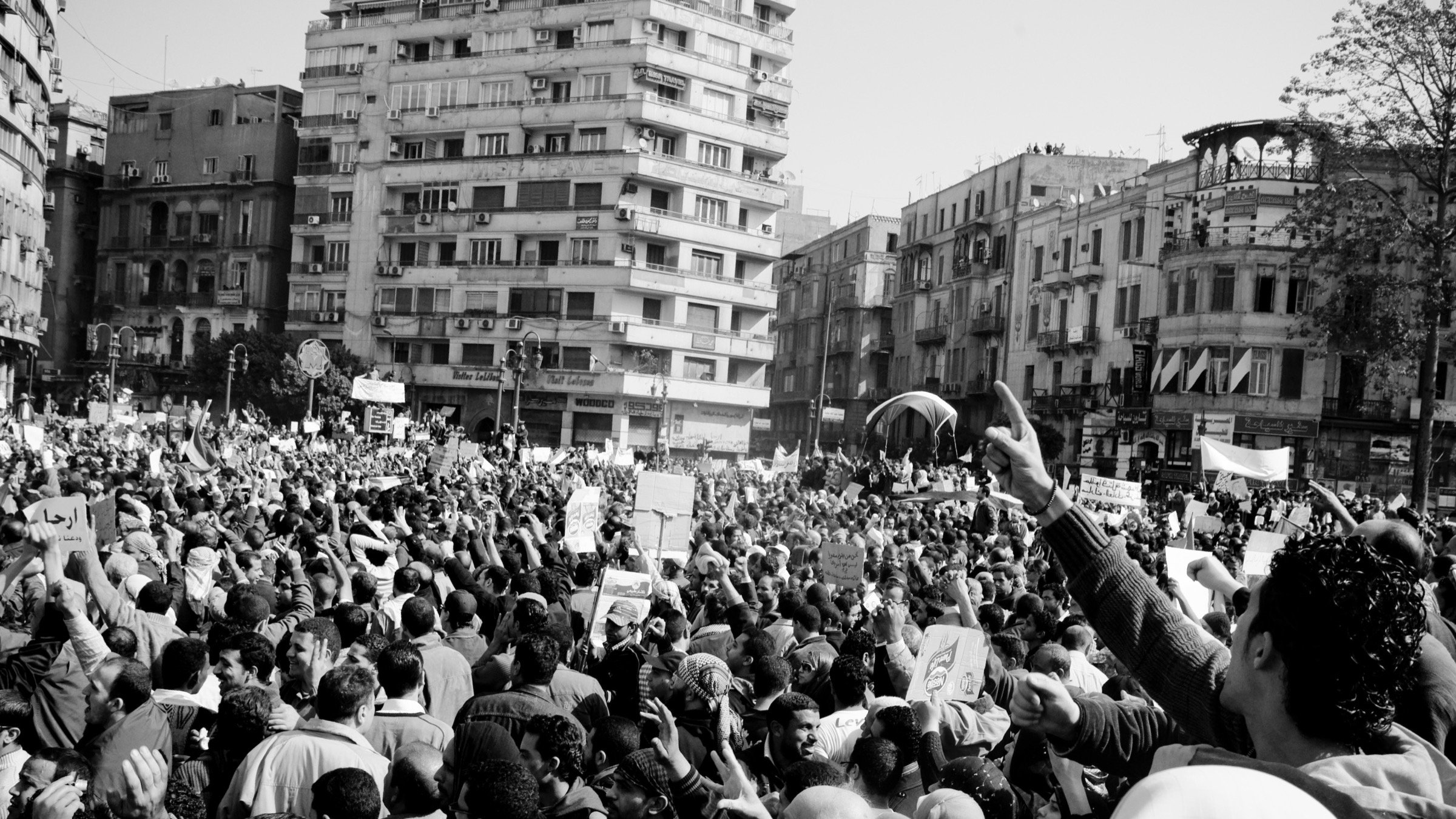 L'image montre une grande foule rassemblée dans une ville, probablement lors d'une manifestation ou d'une protestation. Les participants brandissent des pancartes et semblent exprimer des opinions fortes. L'atmosphère est dynamique et engagée, avec beaucoup de personnes levées les bras, ce qui suggère une ambiance de mobilisation collective. Le noir et blanc de l'image accentue les émotions et l'intensité du moment. Les bâtiments environnants ajoutent un contexte urbain à cette scène de rassemblement.