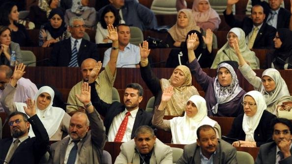L'image montre un rassemblement dans un espace législatif, probablement au sein d'une assemblée ou d'un parlement. De nombreuses personnes sont présentes, avec une majorité levant la main, ce qui indique probablement un vote ou une approbation. Les participants semblent porter des tenues formelles, et certaines femmes portent le hijab. On observe une diversité de participants, tant en termes de genre que d'âge. L'environnement évoque une atmosphère de discussion et de prise de décision collective.