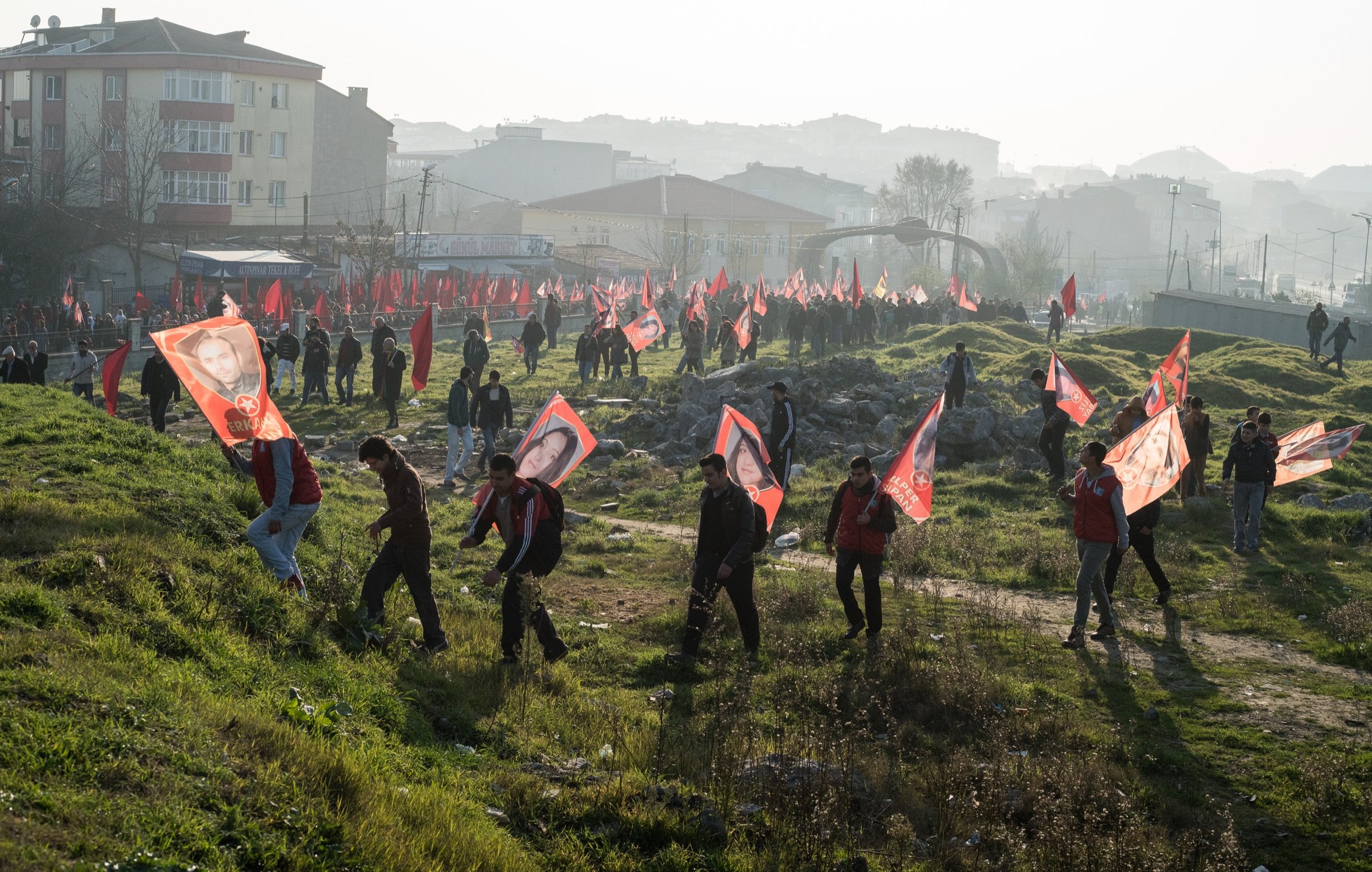 L'image montre un rassemblement de personnes marchant sur un terrain, probablement lors d'une manifestation ou d'une célébration. Les participants portent des drapeaux rouges et des bannières avec des portraits, suggérant un soutien à une cause ou à un leader spécifique. En arrière-plan, on peut voir un paysage urbain flou avec des bâtiments. L'atmosphère semble animée, et la luminosité indique qu'il pourrait s'agir d'une matinée ou d'un après-midi. La présence de nombreux drapeaux rouges donne une impression de rassemblement massif.