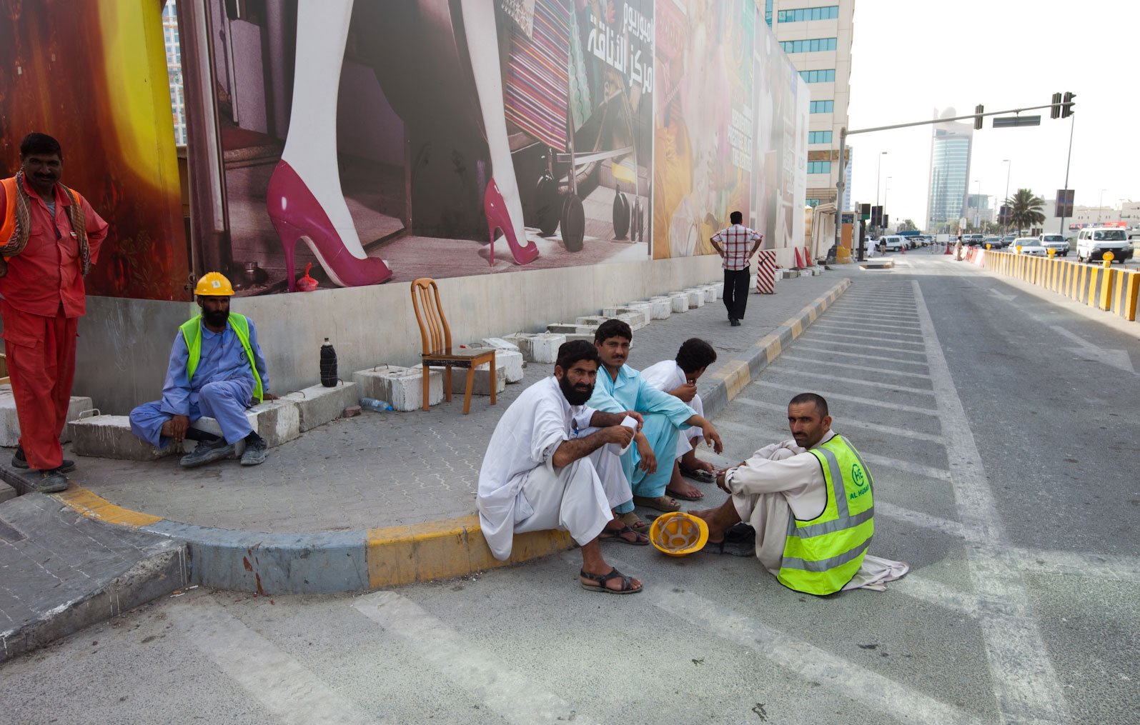 L'image montre un groupe d'hommes assis sur un trottoir, probablement en train de faire une pause. Ils portent des vêtements traditionnels, et certains portent des gilets de travail. En arrière-plan, on aperçoit un grand panneau publicitaire avec des images colorées. L'environnement urbain est visible, avec des bâtiments modernes et une route. L'atmosphère semble calme, et les hommes semblent discuter entre eux.