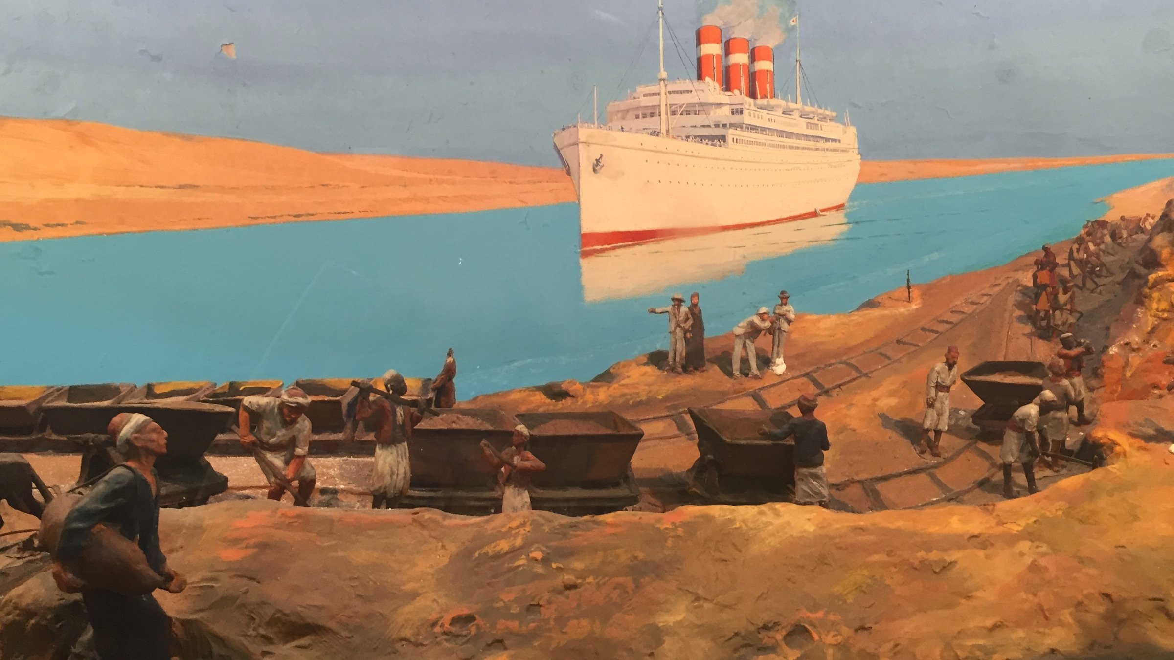 L'image représente une scène historique dans un environnement maritime. On voit un grand paquebot avec trois cheminées rouges amarré près d'une berge, probablement dans un canal ou un fleuve. Au premier plan, plusieurs hommes travaillent sur la rive, transportant des charges, probablement des matériaux ou des marchandises. À l'arrière-plan, le paysage désertique montre des dunes de sable sous un ciel bleu, créant un contraste avec l'eau turquoise. Cette image évoque l'activité maritime et le travail acharné des ouvriers dans un contexte industriel.