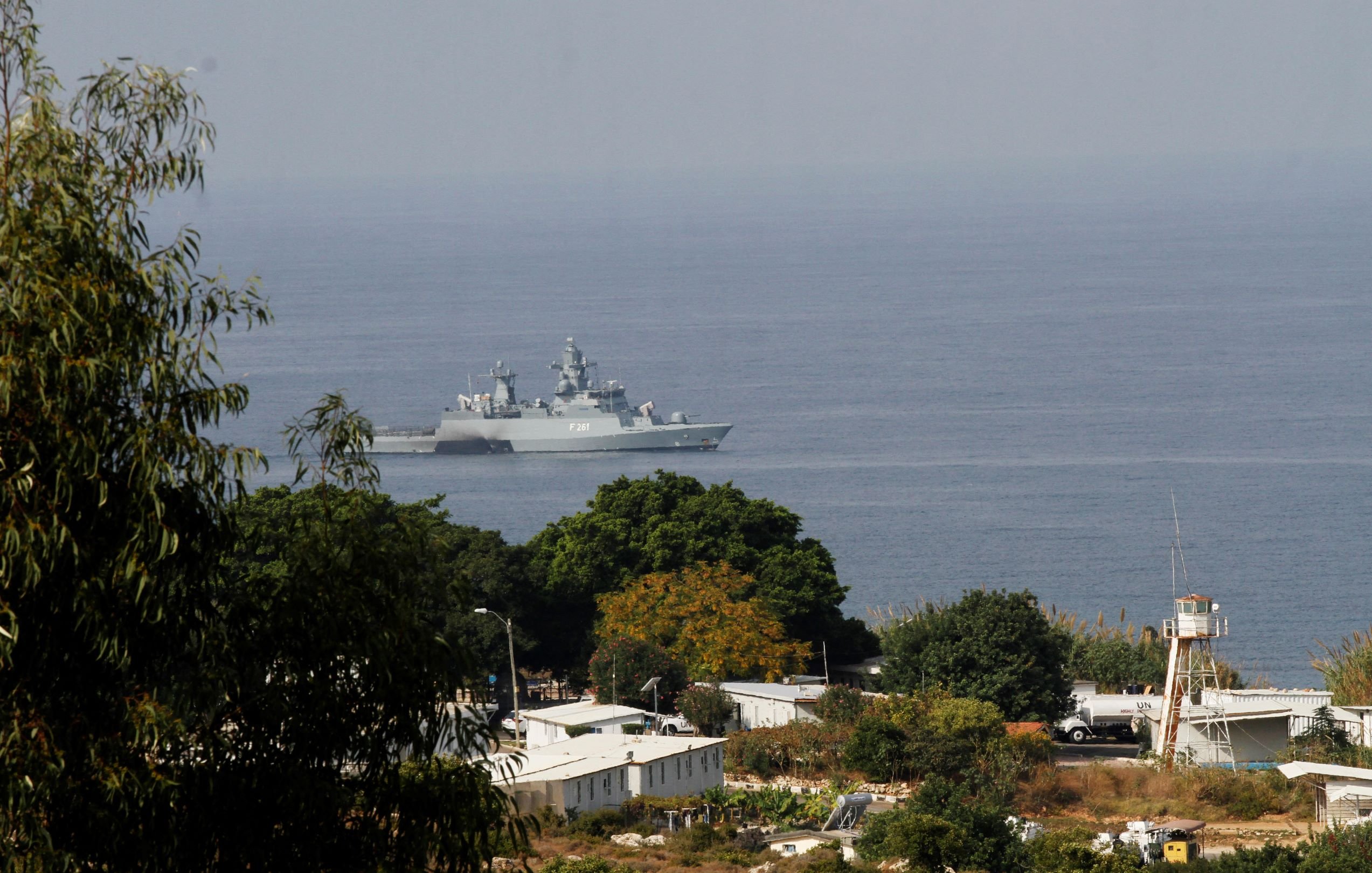 L'image montre un navire militaire se trouvant dans une étendue d'eau, probablement en mer. On aperçoit des paysages côtiers avec quelques arbres et des bâtiments en arrière-plan. Le navire a une structure moderne et est visible au loin, tandis que la végétation au premier plan donne une impression de profondeur à la scène. L'atmosphère semble claire, suggérant une journée ensoleillée.