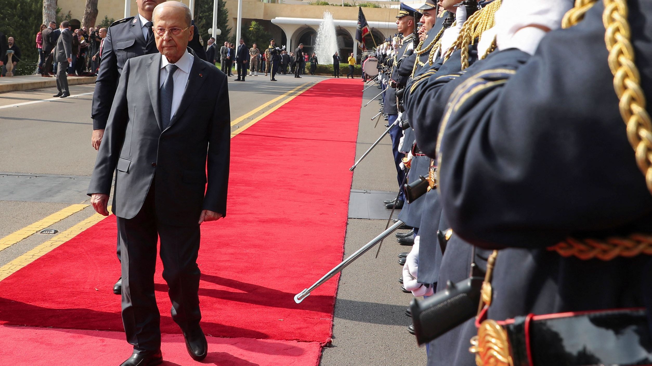 L'image montre une cérémonie officielle. Un homme en costume, probablement une personnalité politique, marche sur un tapis rouge au centre de la scène. De chaque côté, des membres d'une garde d'honneur en uniforme militaire sont alignés, tenant des armes. En arrière-plan, on aperçoit des bâtiments officiels et des drapeaux. L'atmosphère semble solennelle et formelle, suggérant une réception ou un événement diplomatique.
