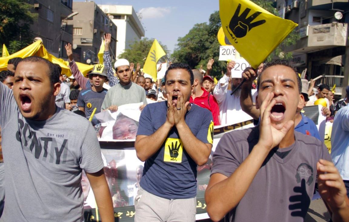 L'image montre une manifestation animée, avec des participants portant des t-shirts colorés. Ils expriment des slogans, certains scandent en levant les bras, tandis que d'autres tiennent des pancartes, notamment avec un symbole en forme de main. L'ambiance semble être pleine d'énergie et de détermination, reflétant une forte mobilisation autour d'une cause. Les bâtiments en arrière-plan suggèrent un milieu urbain.