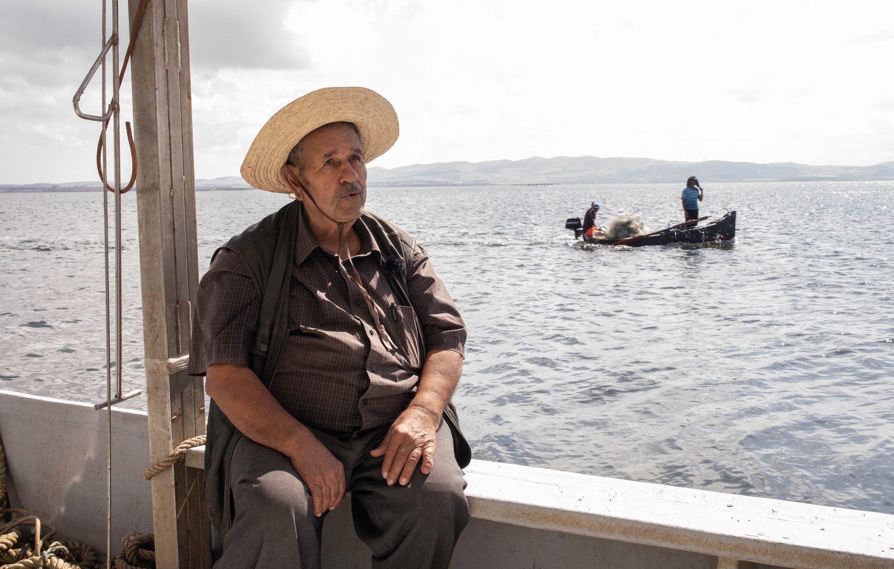 L'image montre un homme assis sur le bord d'un bateau, portant un chapeau de paille large. Il a l'air pensif et regarde vers le large. En arrière-plan, on aperçoit un autre bateau avec des personnes qui semblent s'affairer à la pêche. L'environnement est calme, avec des eaux calmes et un ciel légèrement nuageux, suggérant une ambiance paisible.