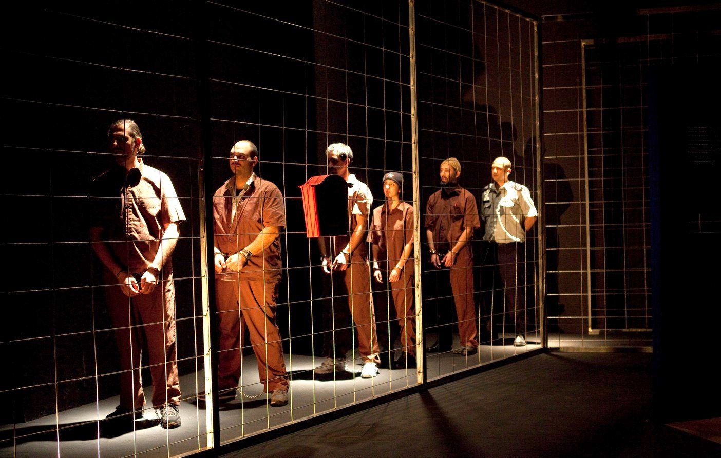 L'image montre un groupe d'hommes disposés en ligne, chacun portant des vêtements de style uniforme. Ils se tiennent derrière un grillage, ce qui crée une impression de séparation ou de détention. L'éclairage est sombre, mettant en valeur les figures des hommes, qui semblent concentrés ou pensifs. L'ambiance générale évoque des thèmes de confinement et d'isolement.