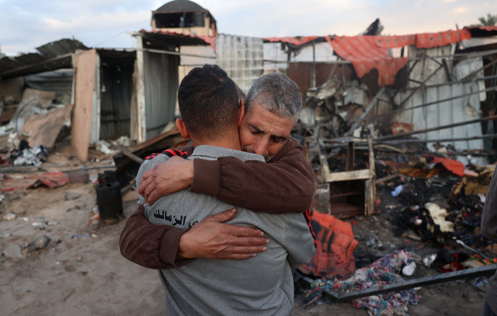 L'image montre deux hommes qui s'étreignent de manière émotive au milieu de décombres. En arrière-plan, il y a des restes de bâtiments détruits, avec des morceaux de métal et des débris éparpillés. L'ambiance semble très chargée d'émotion, suggérant une perte ou un chagrin. Les expressions des hommes témoignent d'une connexion forte et d'un besoin de réconfort dans un moment difficile.