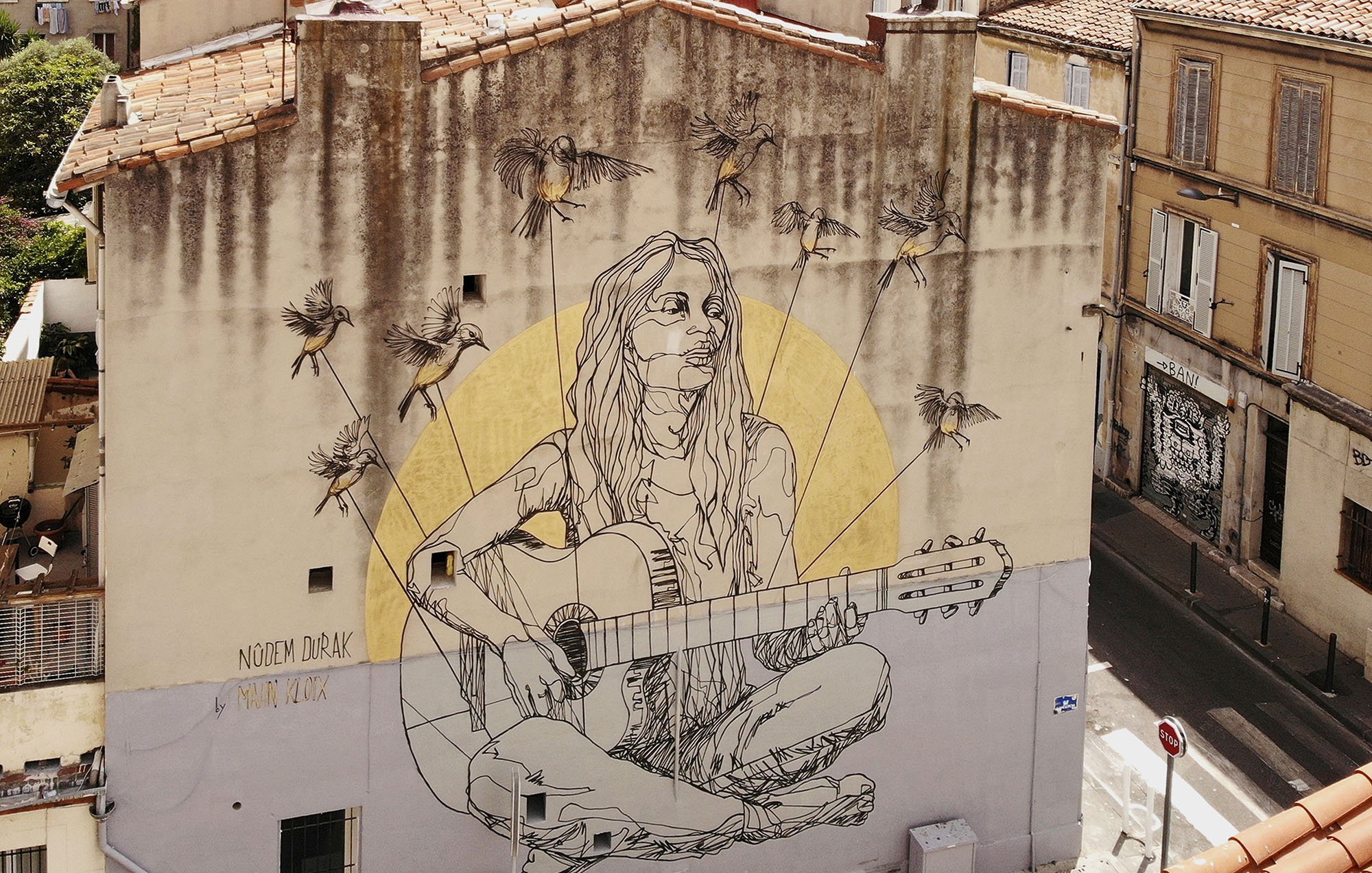L'image montre une fresque murale colorée sur un bâtiment. Au centre, on voit une représentation d'une jeune femme assise, jouant de la guitare. Elle a de longs cheveux et un look décontracté. Autour d'elle, il y a plusieurs abeilles illustrées. En arrière-plan, on aperçoit des bâtiments typiques avec des fenêtres et des toits d'une ville. La fresque semble apporter une ambiance artistique et vivante à l'environnement urbain.