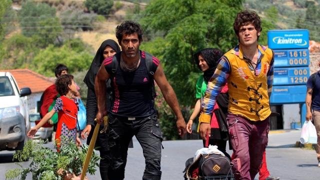 L'image montre un groupe de personnes se déplaçant le long d'une route. Il y a des enfants et des adultes, certains portent des vêtements colorés. Deux hommes avancent avec des paniers à roulettes, contenant probablement des biens ou de la nourriture. L'un d'eux tient une plante, tandis que l'autre semble porter une baguette ou un long objet. On peut voir des maisons et des arbres en arrière-plan, ajoutant une touche de nature à la scène. L'ambiance générale évoque un déplacement ou un voyage.