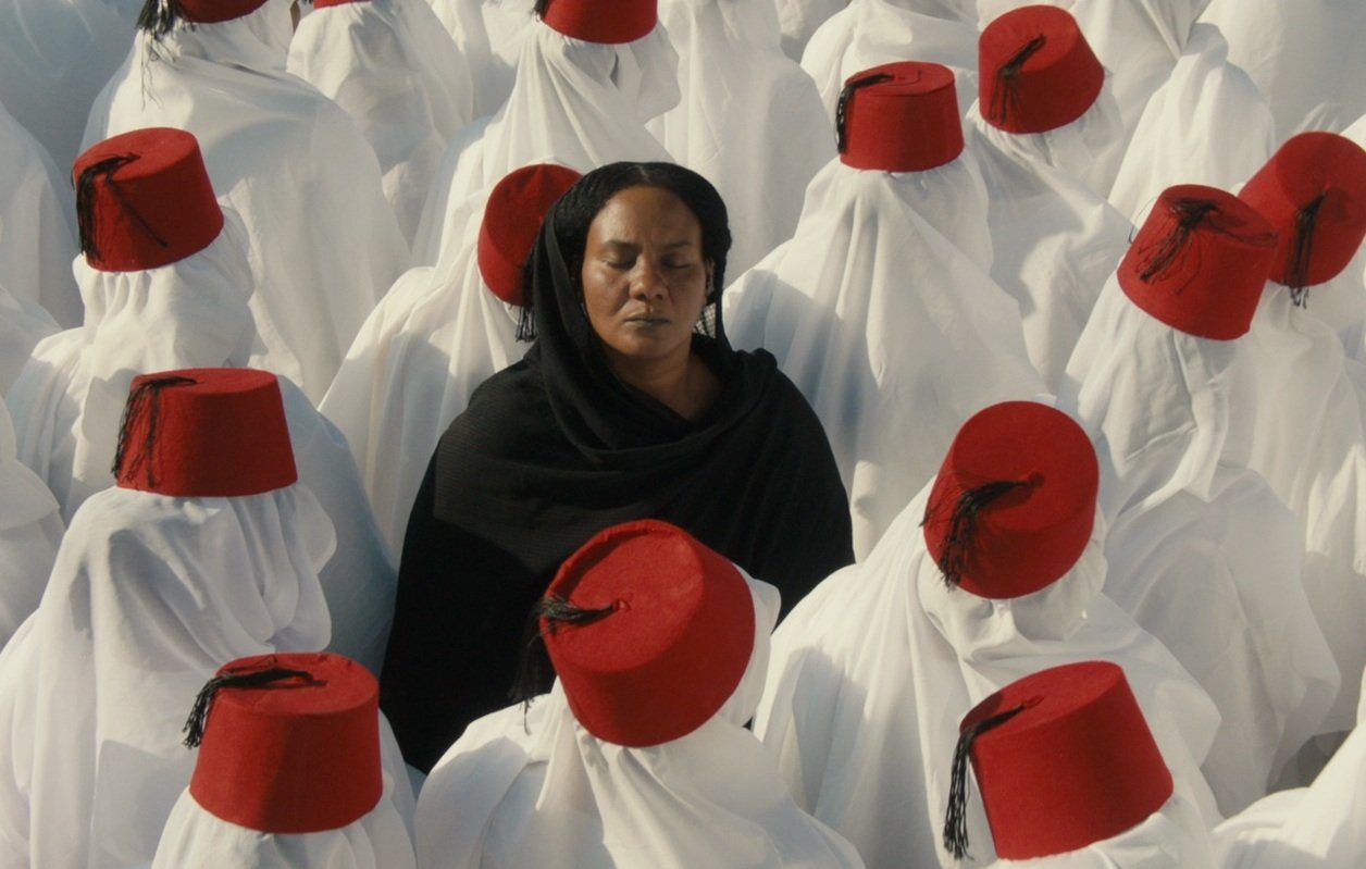L'image montre un groupe de personnes vêtues de longues robes blanches, portant des fez rouges. Au centre, une femme en noir se tient les yeux fermés, contrastant avec l'uniformité des autres. Cette composition crée une atmosphère de méditation ou de réflexion, avec un fort accent sur la spiritualité et la communauté. Les couleurs et les vêtements évoquent une tradition culturelle riche.
