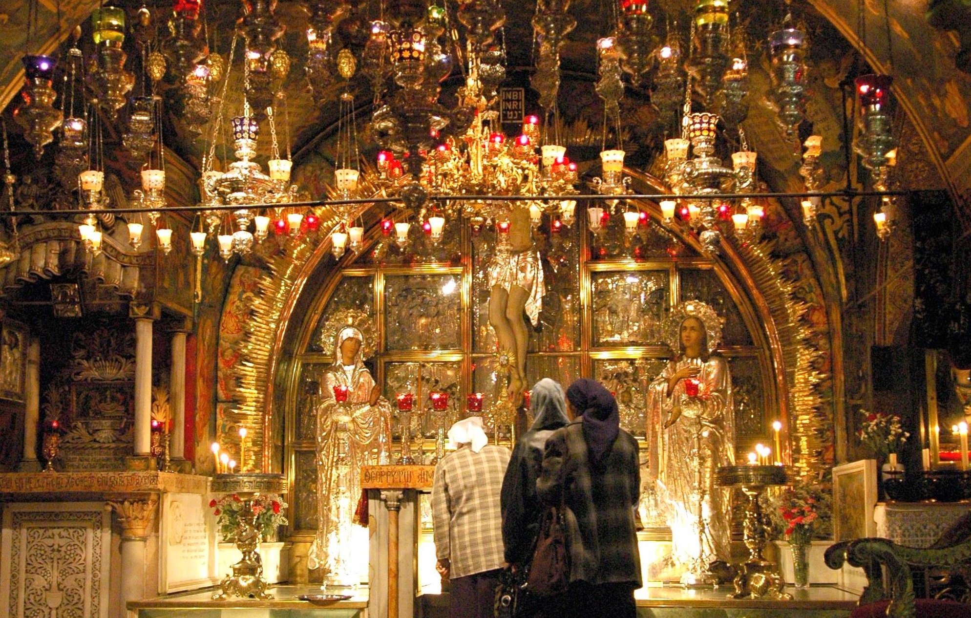 L'image montre l'intérieur d'un lieu de culte, probablement une église ou une chapelle. L'architecture est ornée, avec un plafond voûté richement décoré. On peut voir des lumières colorées suspendues et des candélabres, créant une ambiance chaleureuse et mystique. Au centre, il y a des figures dorées, possiblement des icônes ou des statues, entourées de bougies et de fleurs. Quelques personnes, vêtues de manteaux et de foulards, se tiennent devant cette scène, semblant s'engager dans une prière ou une contemplation.