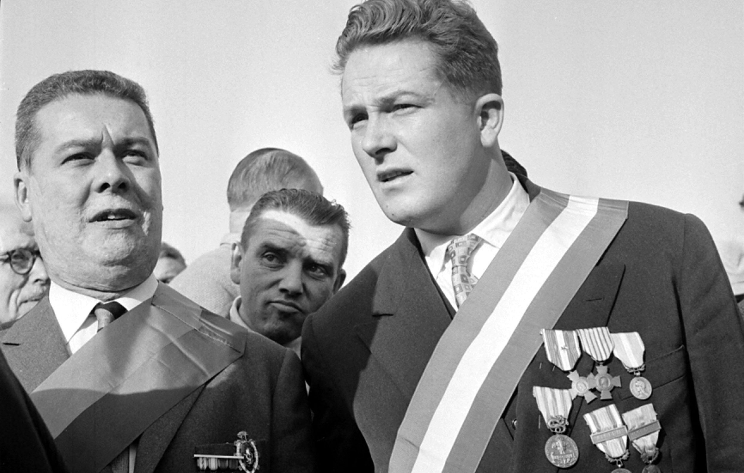 L'image montre deux hommes qui semblent participer à un événement officiel ou une cérémonie. L'un des hommes porte plusieurs médailles accrochées à sa veste, ainsi qu'une écharpe. L'autre homme porte également une écharpe, mais il a moins de médailles visibles. L'ambiance paraît solennelle, avec d'autres personnes en arrière-plan. Les expressions des deux hommes suggèrent qu'ils sont engagés dans une conversation ou qu'ils se concentrent sur l'événement.