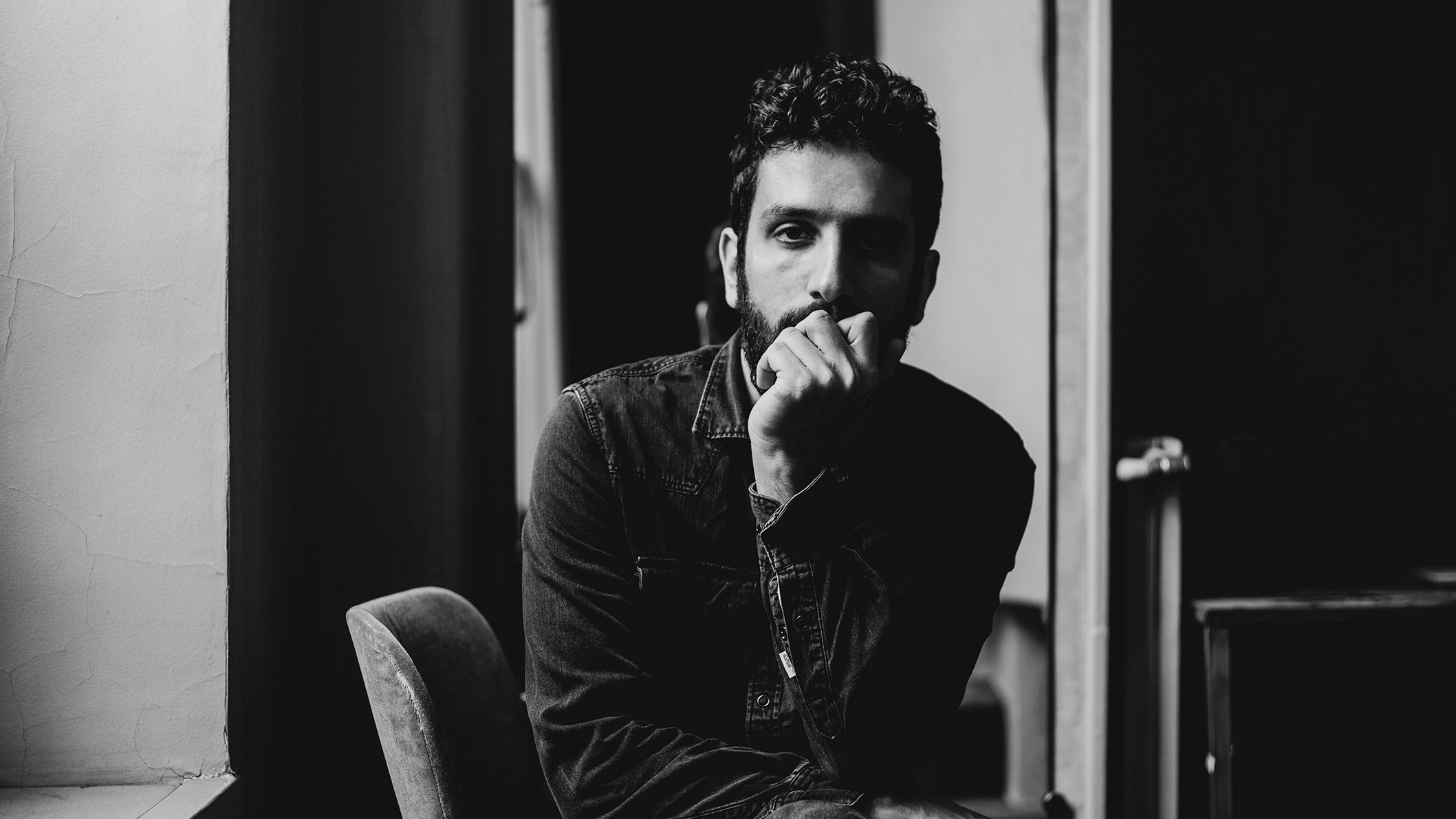 L'image montre un homme assis sur une chaise dans un intérieur sombre. Il a une barbe et porte une chemise en denim. Son expression est réfléchie, avec une main sur le menton, tandis qu'il regarde en direction de la caméra. L'éclairage met en valeur les contours de son visage et crée une ambiance introspective. Des éléments de l'environnement, comme une fenêtre et un miroir, ajoutent de la profondeur à la composition.