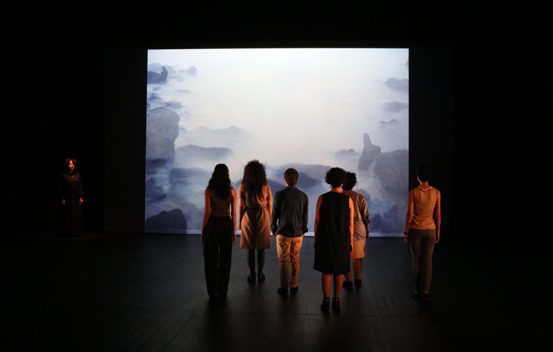 L'image montre une scène de performance artistique. On y voit plusieurs personnes de dos, regardant un grand écran qui projette une image évoquant un paysage marin brumeux. En arrière-plan, on aperçoit une silhouette d'une personne habillée en noir, se tenant à gauche de l'image. Les autres participants, vêtus de différentes tenues, semblent absorbés par la projection. L'éclairage est tamisé, créant une ambiance contemplative.