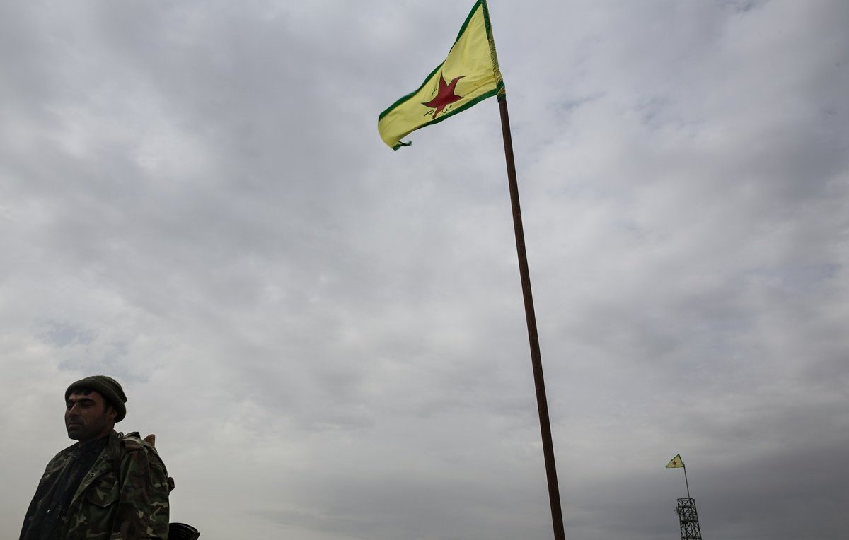 L'image montre un homme en uniforme militaire se tenant près d'un drapeau. Le drapeau est de couleur jaune avec une étoile rouge au centre, entouré de bandes vertes. L'arrière-plan est nuageux, ce qui donne un ton sombre à la scène. En plus de l'homme, on peut apercevoir une autre instance du drapeau en arrière-plan, également hissé sur un mât.