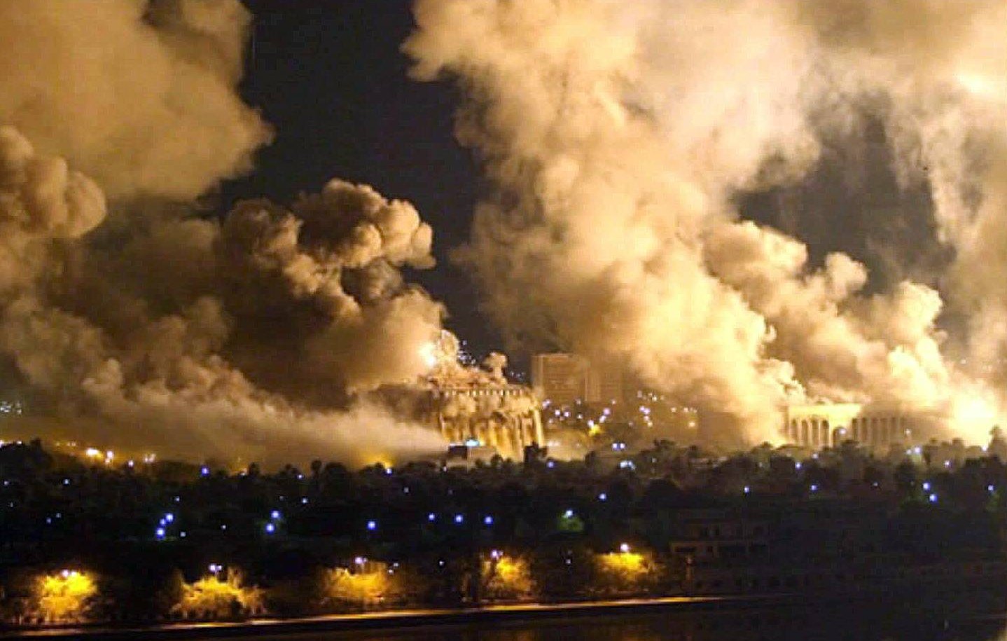 L'image montre une scène nocturne dramatique avec des nuages de fumée épais s'élevant vers le ciel. On peut voir des structures lumineuses en arrière-plan, suggérant que des bâtiments sont présents, mais ils sont partiellement obscurcis par la fumée. La réflexion des lumières sur une surface d'eau au premier plan ajoute une atmosphère sombre et intense à la composition. L'ensemble évoque une situation de conflit ou de désastre.