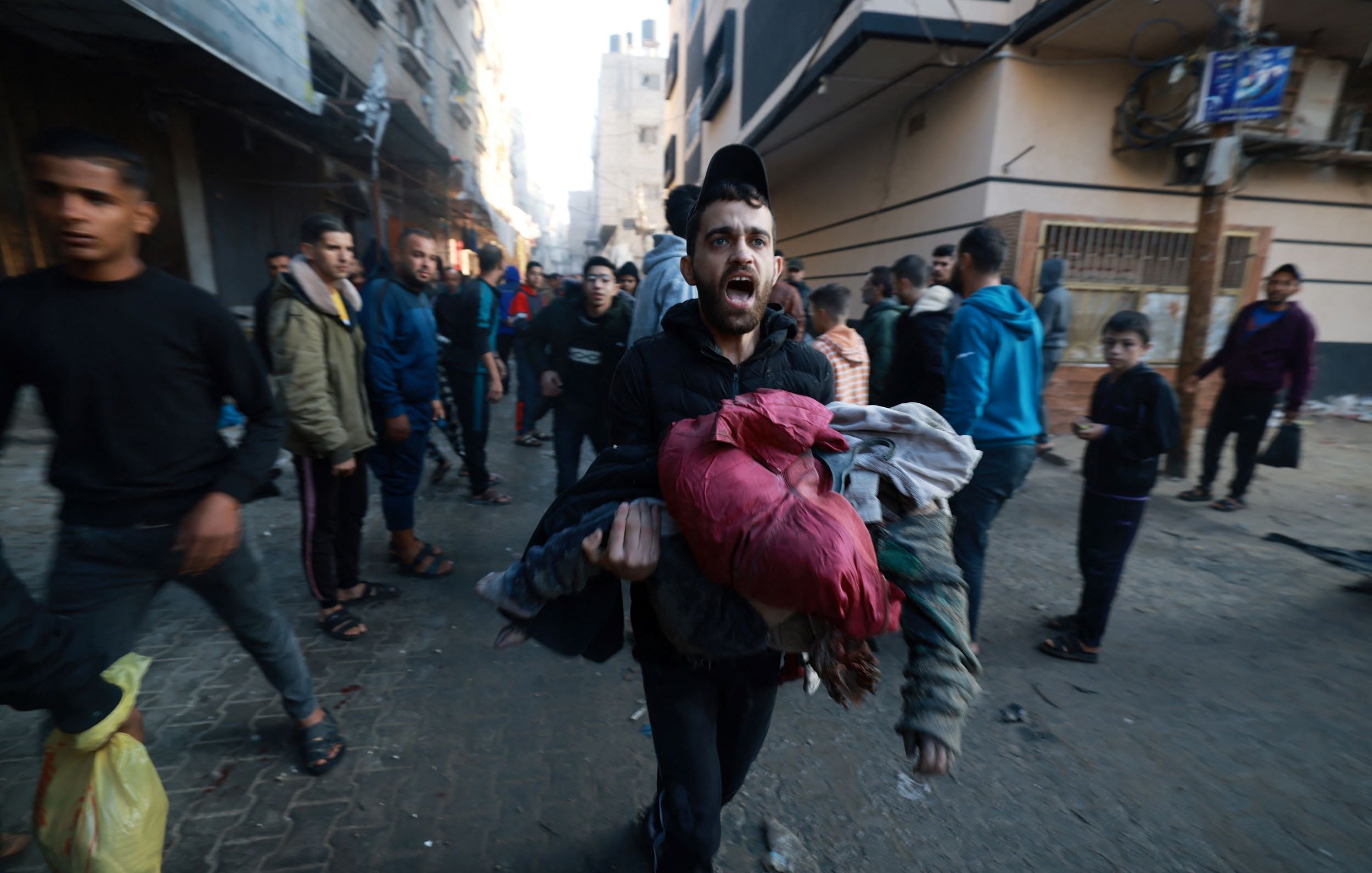 L'image montre une scène urbaine animée où un homme, visiblement en détresse, transporte des vêtements ou des objets dans ses bras. Il semble crier ou exprimer une émotion forte, tandis que d'autres personnes marchent autour de lui. L'environnement est urbain, avec des bâtiments en arrière-plan qui semblent endommagés ou en délabrement. L'atmosphère de l'image suggère une situation de crise ou d'urgence, avec une foule qui se déplace rapidement.