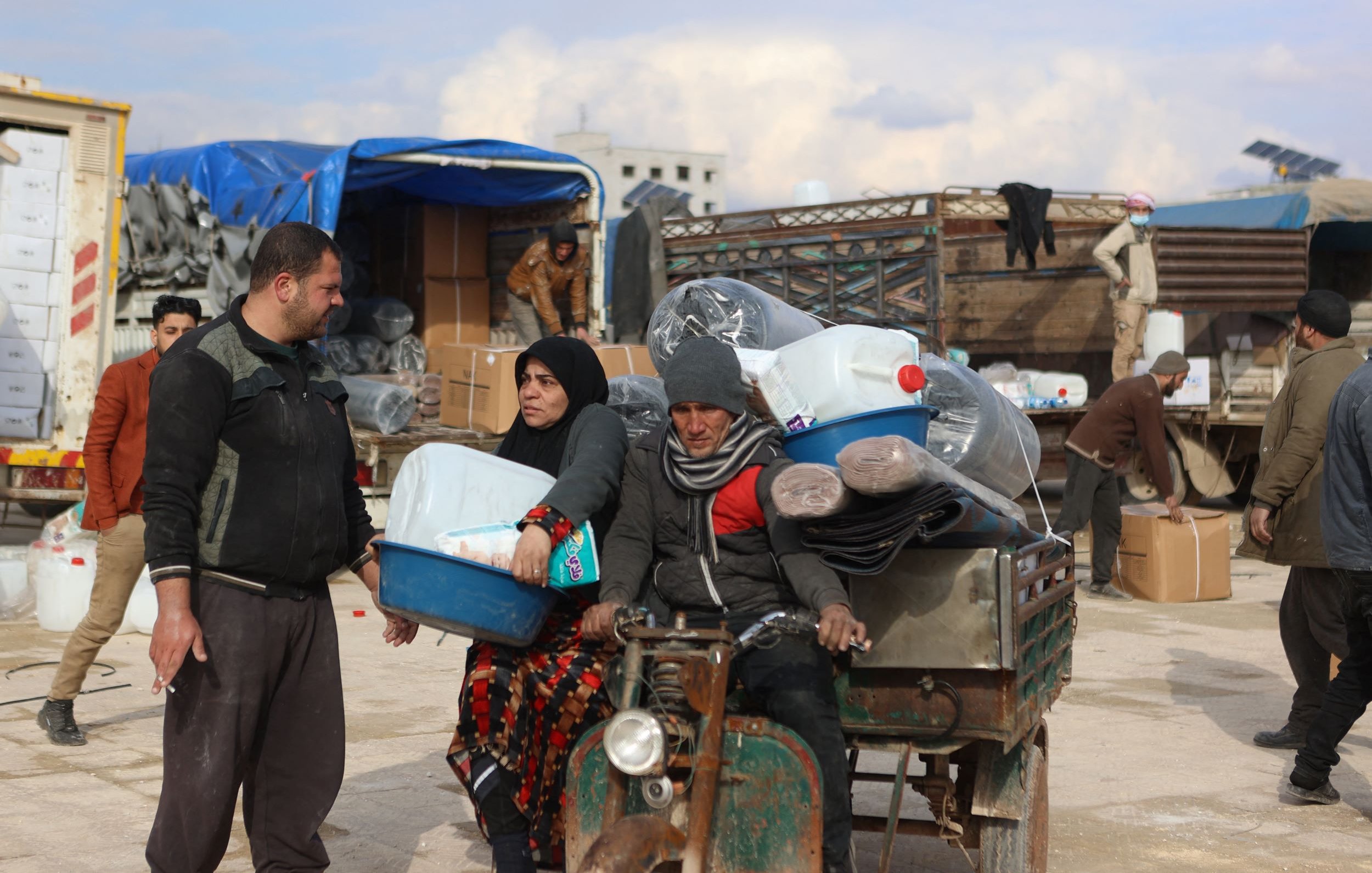 L'image montre une scène de marché ou de distribution dans un environnement rural. On y voit un homme et une femme discutant, tandis qu'un autre homme est assis sur un véhicule à trois roues, probablement en train de transporter des marchandises. Le décor entoure des containers en plastique, et plusieurs personnes s'affairent à décharger ou à déplacer des articles. Le ciel est nuageux, suggérant une atmosphère d'activité et d'échange.