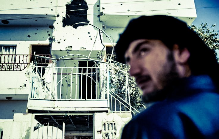 L'image montre un bâtiment portant des marques visibles d'impacts de balles, suggérant des dommages dus à des conflits. Au premier plan, un homme est partiellement visible, tourné vers l'extérieur, avec une expression pensive. L'environnement semble urbain, et le contraste entre le visage de l'homme et le bâtiment endommagé crée une atmosphère de tension et de tristesse.