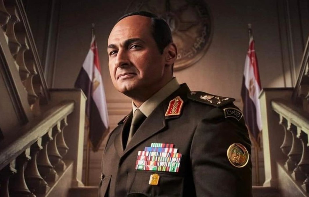 L'image montre un homme en uniforme militaire, vraisemblablement un officier supérieur. Il porte plusieurs décorations et insignes sur sa tenue, ce qui indique un haut rang. En arrière-plan, on aperçoit une architecture grandiose avec des drapeaux, ce qui suggère un environnement officiel, peut-être un gouvernement ou une institution militaire. L'expression de l'homme est sérieuse et déterminée.