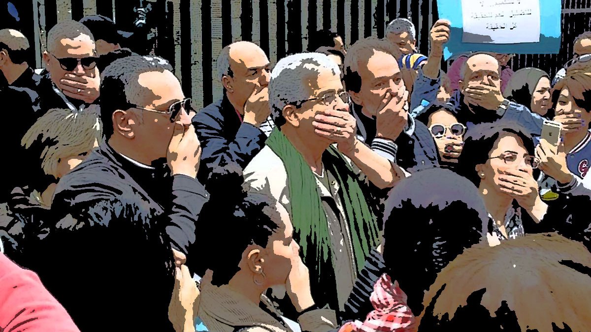 L'image montre un groupe de personnes réunies dans un cadre extérieur. Beaucoup d'entre elles se tiennent avec la main sur la bouche, semblant exprimer un silence ou protester. En arrière-plan, on aperçoit un panneau qui semble contenir un message. L'ambiance générale semble sérieuse et engagée, suggérant une forme de mobilisation ou de manifestation. Les participants semblent concentrés et solidaires.