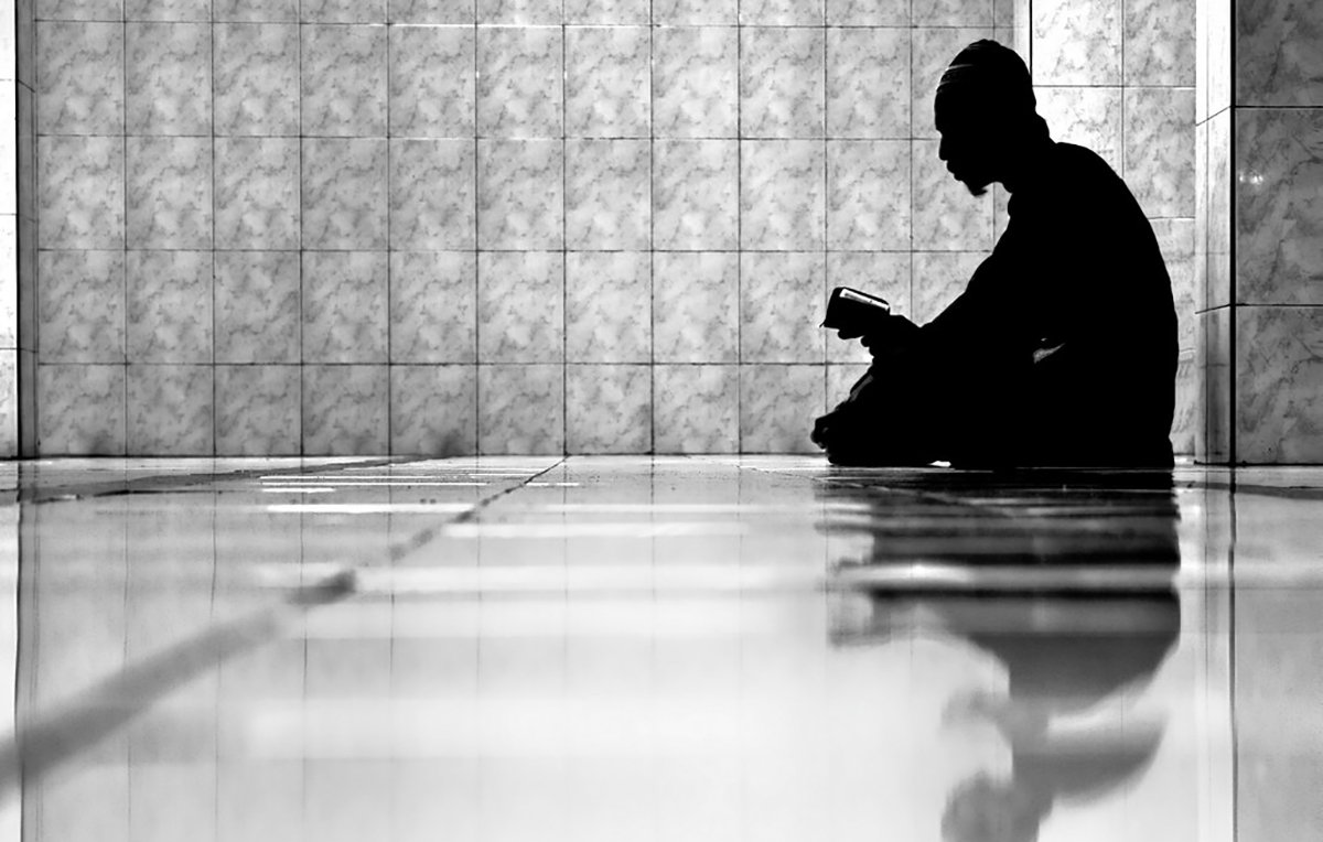 L'image montre un individu assis sur le sol, plongé dans la lecture d'un livre. La composition est en noir et blanc, créant un contraste fort entre l'ombre de la personne et l'éclat des carreaux brillants au sol. L'ambiance est calme et contemplative, et le reflet de la silhouette sur le sol ajoute une dimension introspective à la scène. Les murs en arrière-plan sont recouverts de carrelage, ce qui renforce l'aspect minimaliste de l'image.