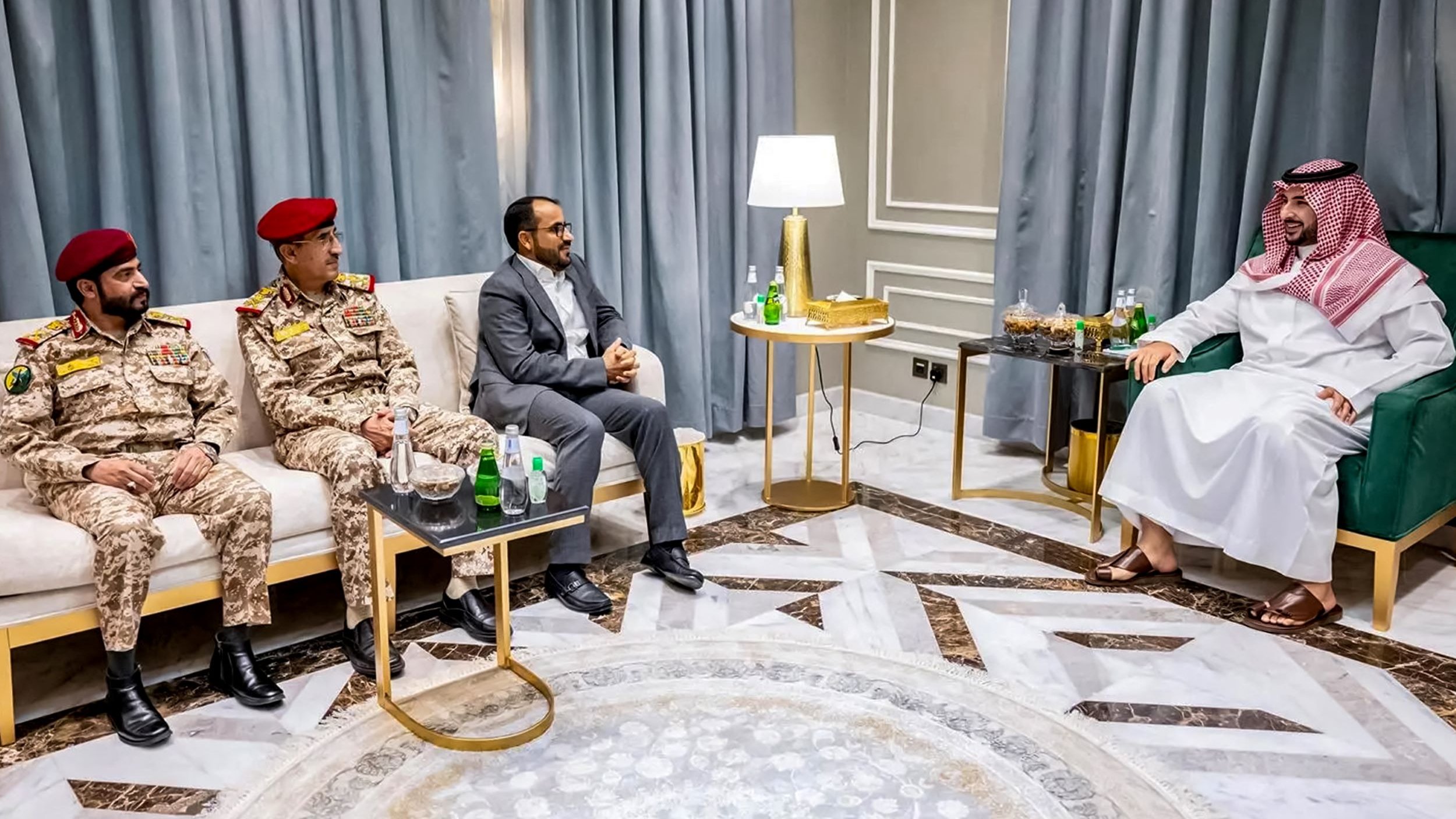 L'image montre une rencontre entre plusieurs hommes assis dans une pièce bien décorée. Deux hommes portent des uniformes militaires, avec des chapeaux rouges, tandis qu'un troisième homme est habillé en costume. Le quatrième homme, à droite, porte une robe traditionnelle. Ils sont assis autour de tables basses où se trouvent des bouteilles d'eau et des verres. Le décor comprend des rideaux gris et un sol en marbre. L'atmosphère semble être celle d'une discussion formelle.