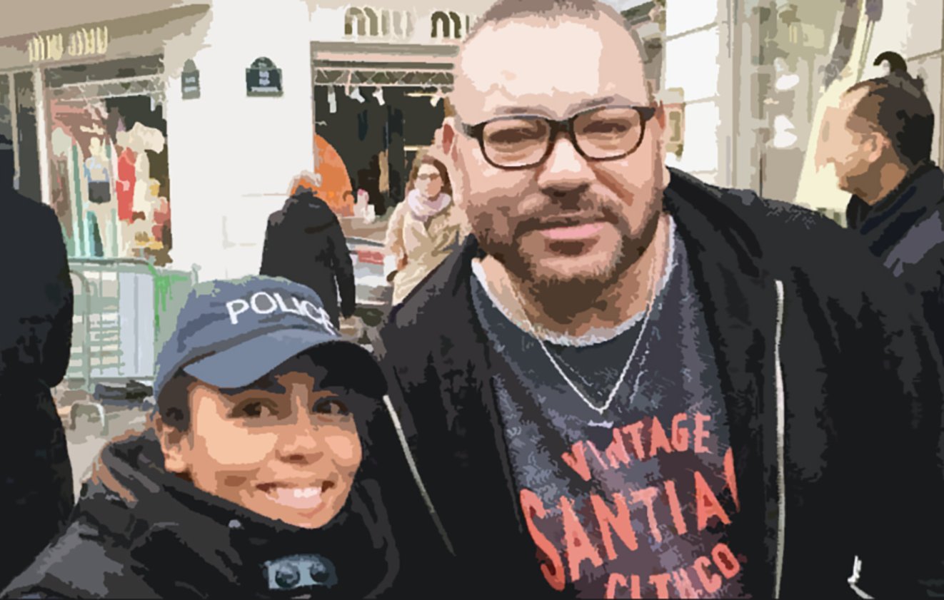 L'image montre deux personnes posant ensemble à l'extérieur. L'une porte une casquette avec le mot "POLICE" et a un sourire, tandis que l'autre, à la barbe bien taillée et portant des lunettes, a une expression amicale. L'arrière-plan montre une zone commerçante animée avec des gens et des boutiques.