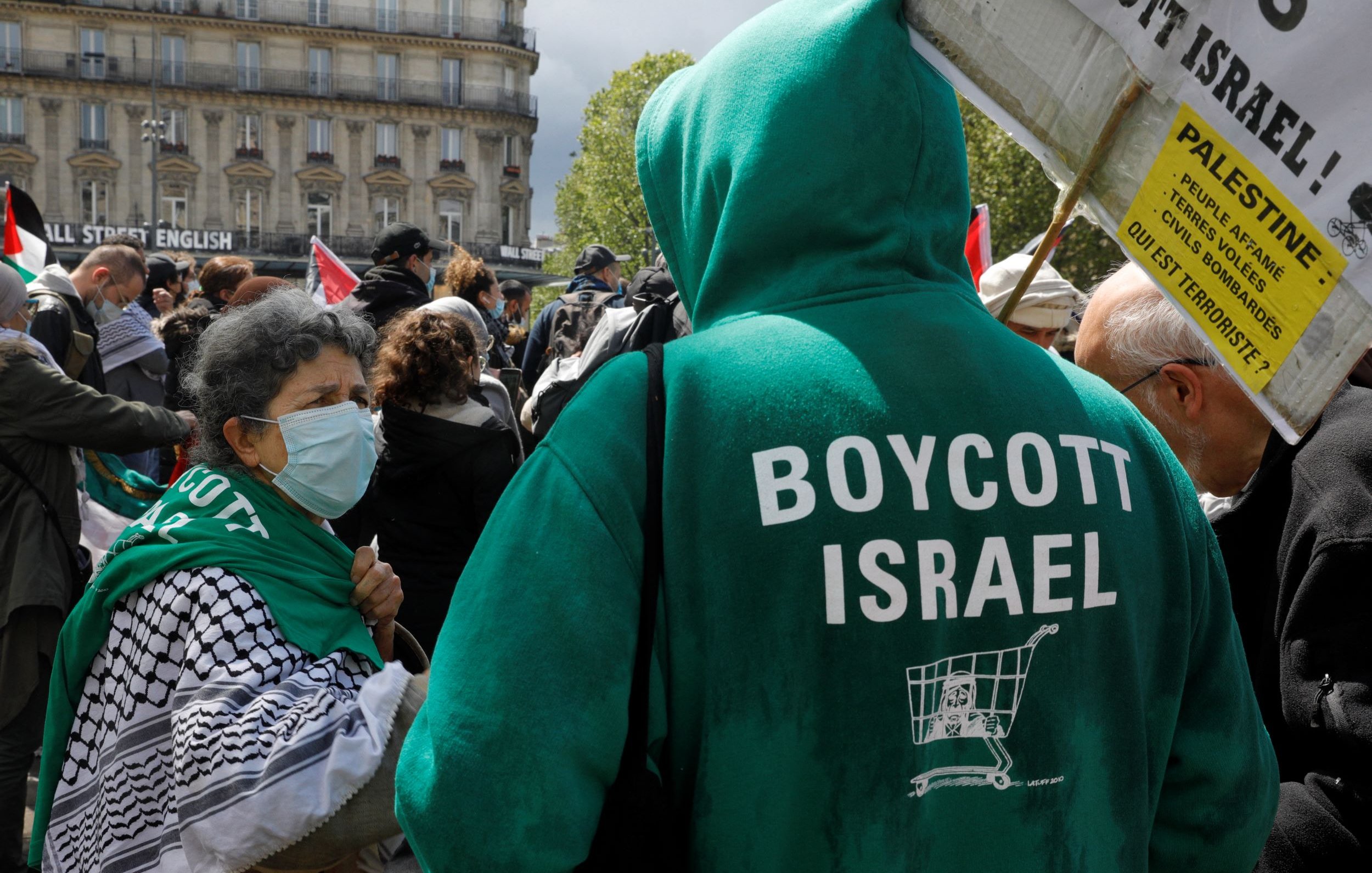 L'image montre une manifestation où des personnes se rassemblent pour exprimer leur soutien à la Palestine. Au premier plan, on voit une femme portant un foulard et un masque, conversant avec une autre personne. Celle-ci porte un sweatshirt vert sur lequel est inscrit "BOYCOTT ISRAEL". En arrière-plan, on distingue plusieurs personnes et des drapeaux, suggérant une mobilisation collective autour de cette cause. L'ambiance semble dynamique et engagée.