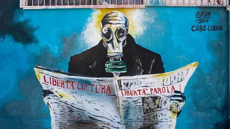 L'image montre une œuvre de street art représentant une figure portant un masque à gaz, lisant un journal. La figure est peinte avec des couleurs vives, créant un contraste avec l'arrière-plan. Sur le journal, il y a des inscriptions en italien, telles que "LIBERTÀ CULTURA" et "LIBERTÀ PAROLA", qui signifient "Liberté de la culture" et "Liberté d'expression". L'ensemble de l'œuvre véhicule un message fort sur la liberté et la société.