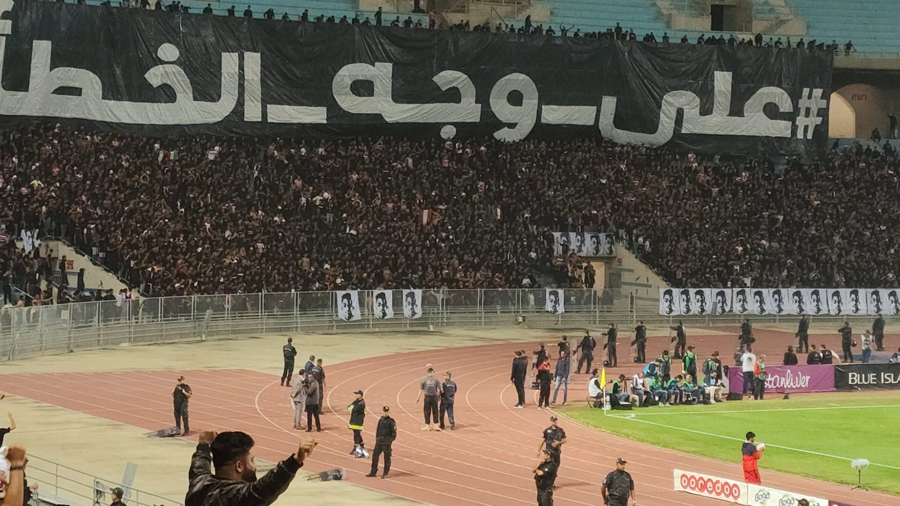 Stade plein de supporters en noir avec une grande banderole en arabe. Énergie et passion.