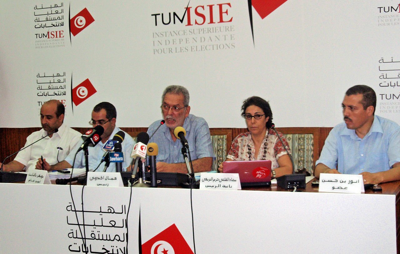 L'image montre une conférence de presse avec plusieurs personnes assises à une table. Un homme au centre semble parler, tandis que les autres personnes à ses côtés écoutent ou prennent des notes. Il y a des microphones sur la table et un arrière-plan avec des logos et du texte en arabe et en français, probablement lié à une institution électorale. L'environnement semble formel et consacré aux affaires publiques.