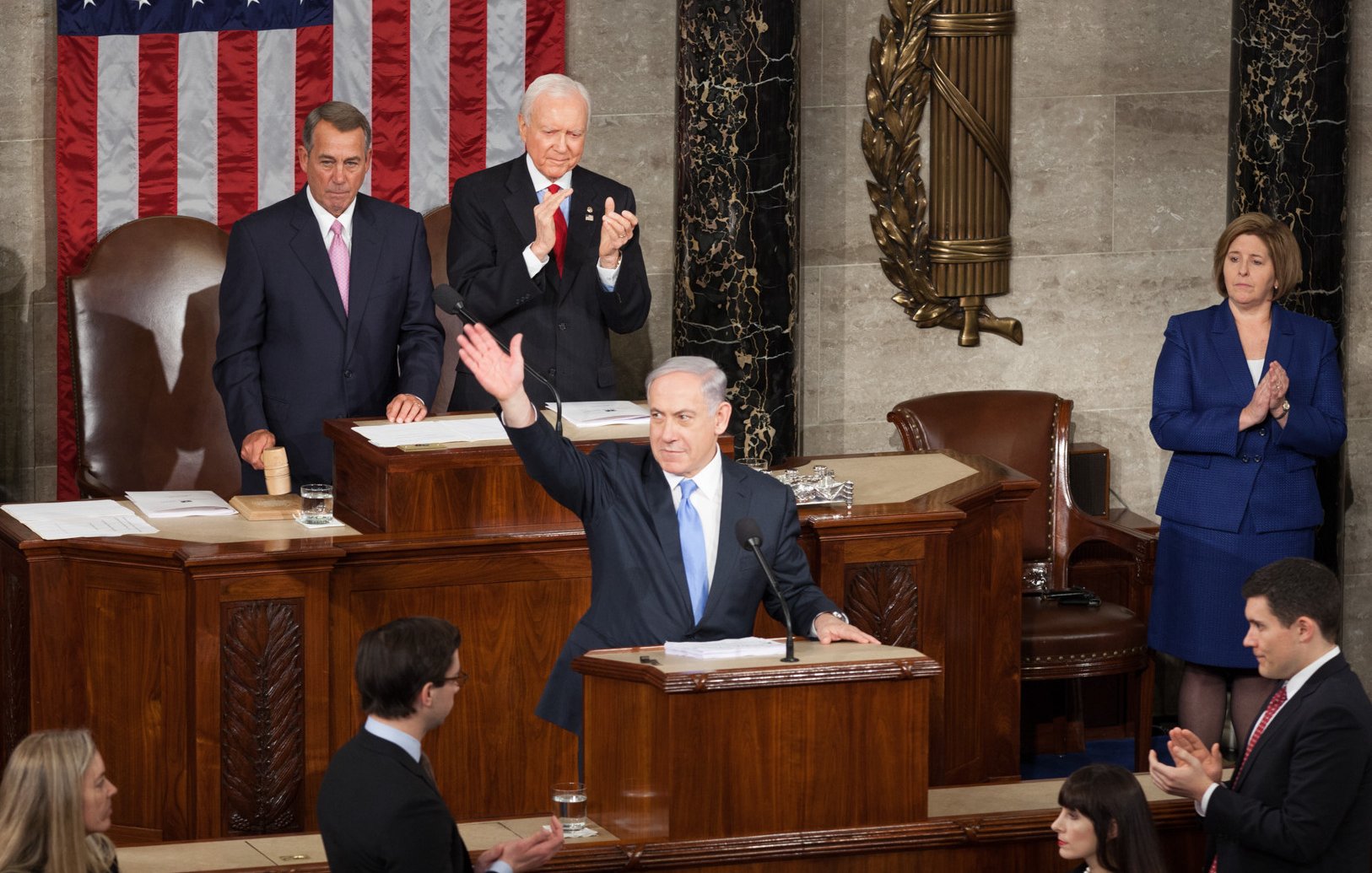 Cette image montre une scène dans une salle législative. Un homme se tient au pupitre, levant la main en signe de salut ou d'allocution. Derrière lui, plusieurs personnes, dont un homme plus âgé et une femme, l'écoutent attentivement. On peut également voir un drapeau américain en arrière-plan, ainsi que le mobilier typique d'une assemblée. L'atmosphère semble formelle et solennelle, suggérant un moment important, potentiellement un discours ou une présentation officielle.