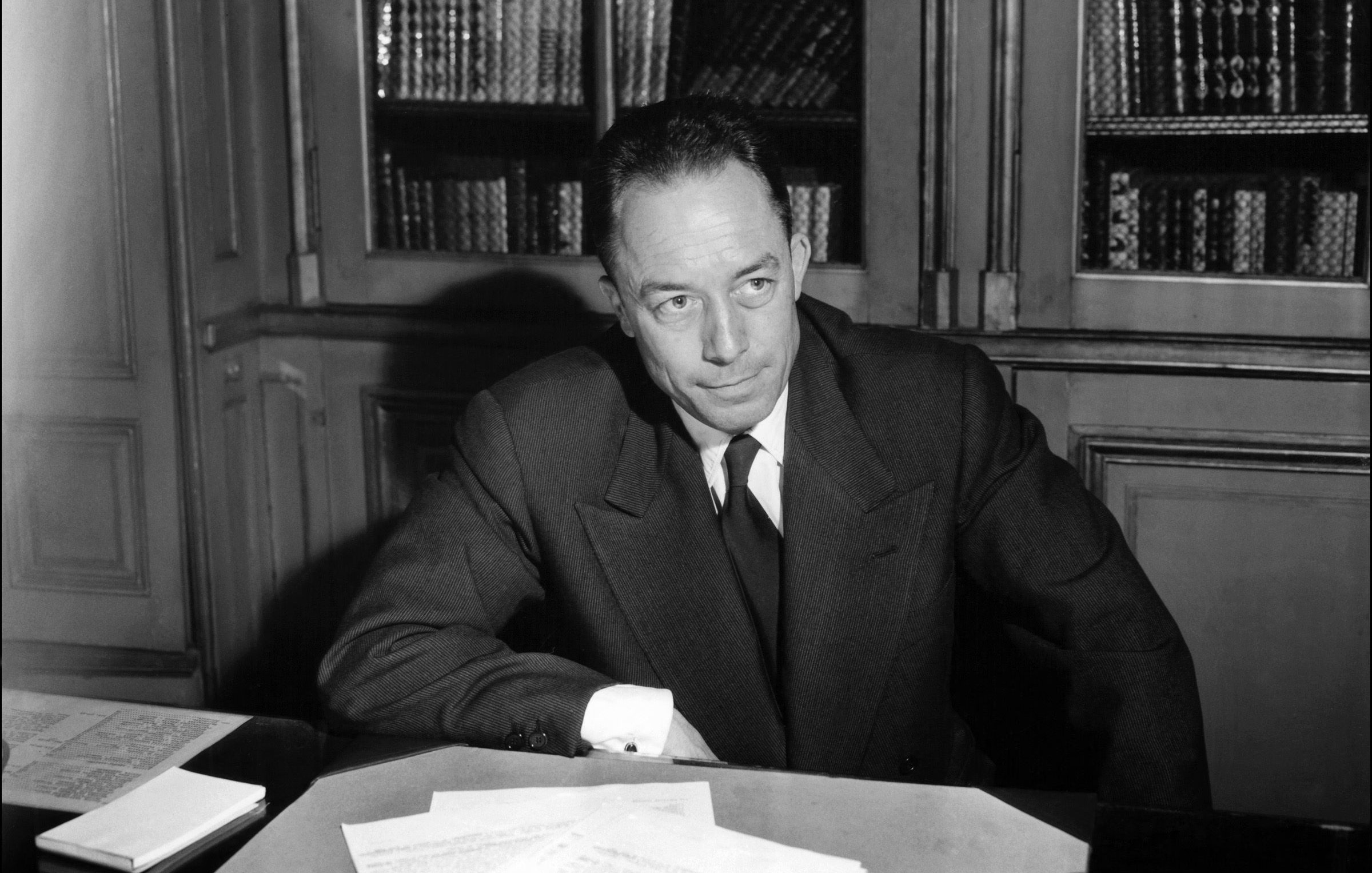 L'image montre un homme assis à un bureau, avec des documents éparpillés devant lui. Il est habillé de manière formelle, portant un costume sombre et une cravate. L'arrière-plan présente des étagères remplies de livres. L'éclairage met en valeur ses traits, suggérant un moment de réflexion ou de concentration.