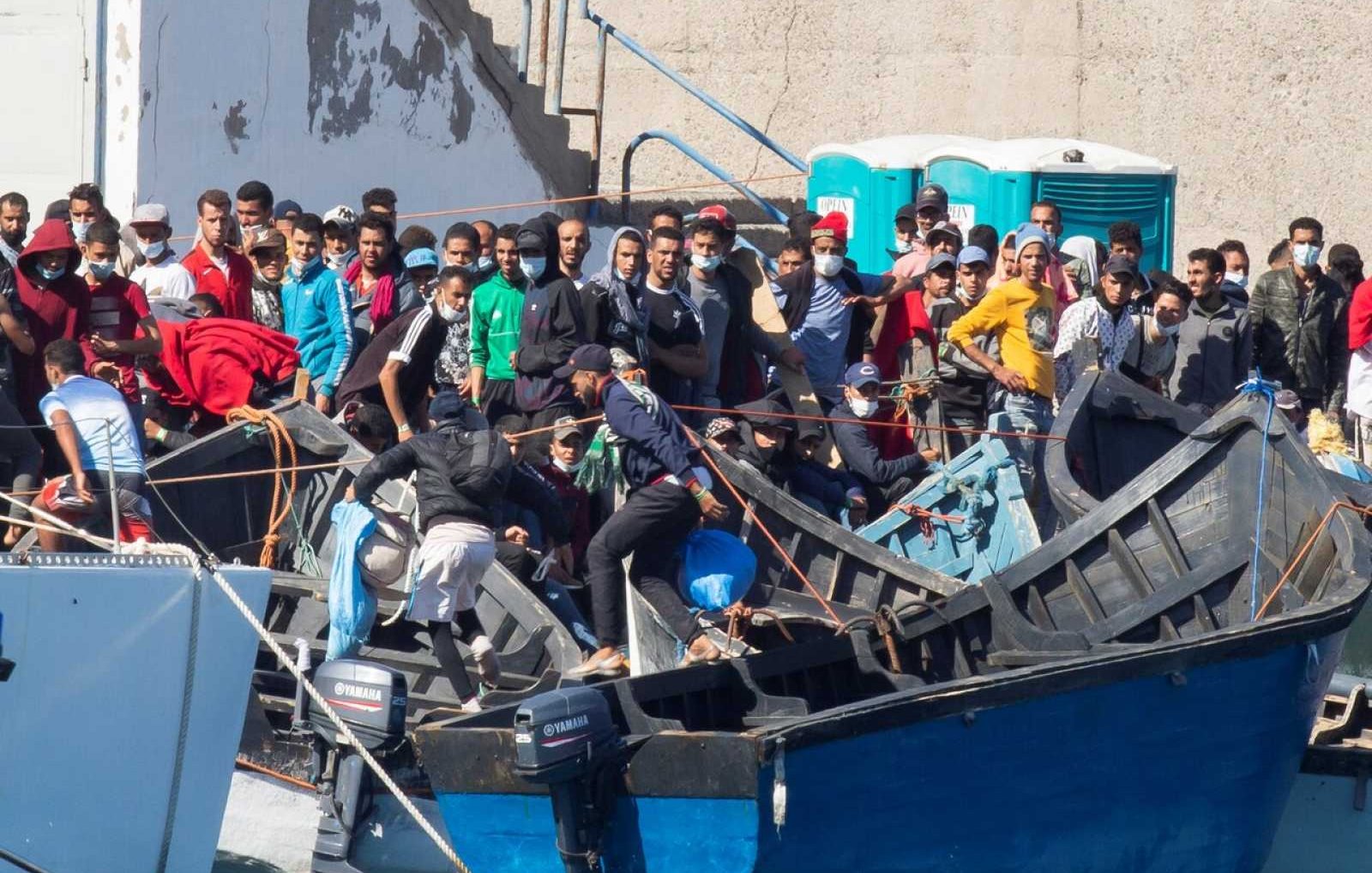 L'image montre un groupe de personnes sur des embarcations, probablement dans un port ou une zone côtière. Les individus semblent rassemblés autour de plusieurs bateaux, avec une diversité de vêtements, certains portant des masques. L'environnement est urbain, et il y a des structures en béton visibles en arrière-plan. On peut noter une atmosphère de mouvement et d'activité parmi les personnes présentes sur les bateaux.