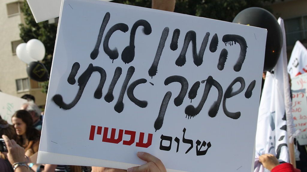L'image montre une personne tenant une pancarte pendant une manifestation. La pancarte est écrite en hébreu, et on peut voir des slogans ou des messages gravés dessus. En arrière-plan, il y a d'autres manifestants et des ballons, ce qui suggère un rassemblement public. L'ambiance semble être celle d'une protestation, avec une atmosphère engagée.