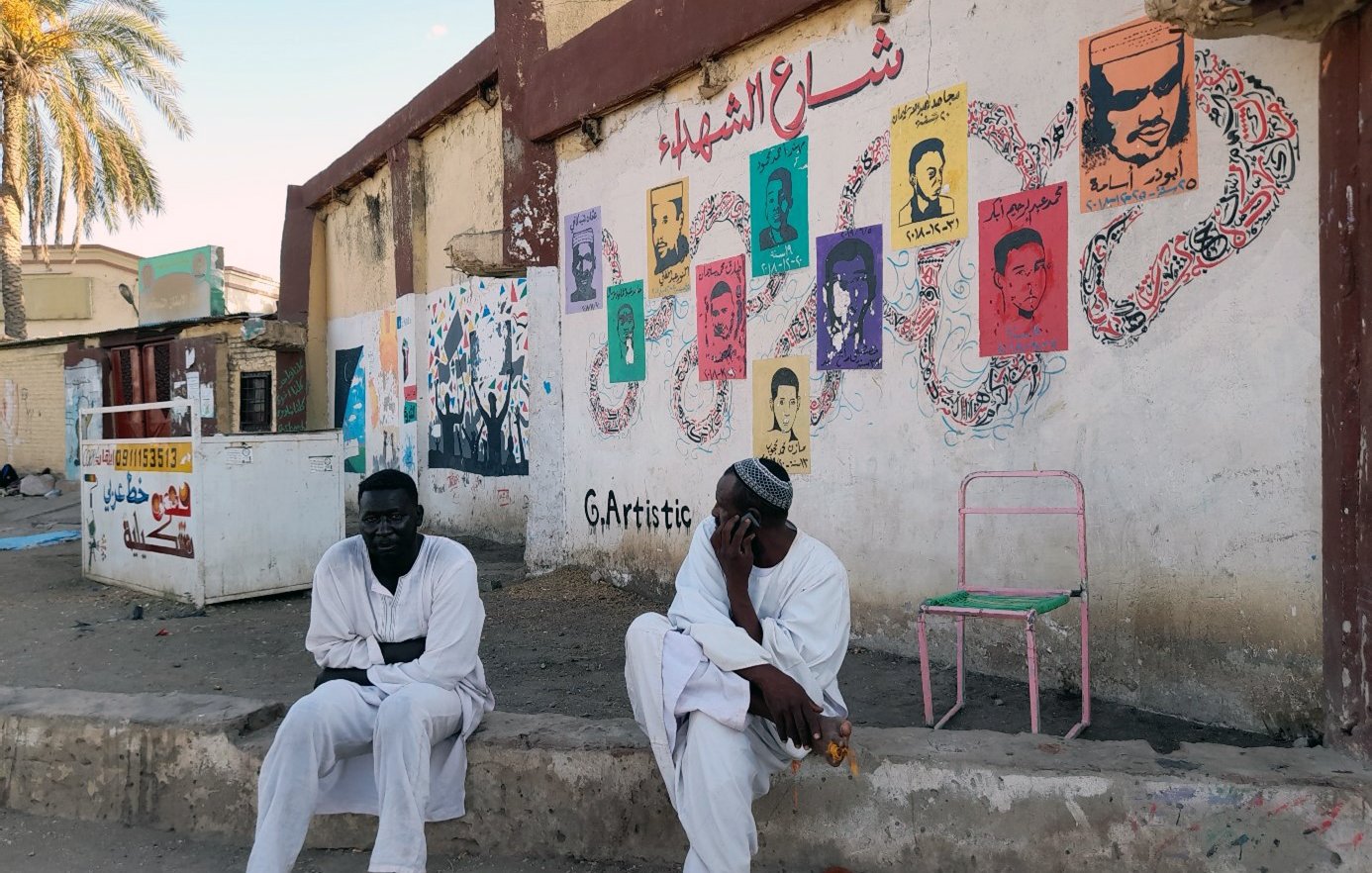 L'image montre deux hommes assis à l'extérieur, probablement dans une rue d'une ville. Ils portent des vêtements traditionnels, en particulier des robes longues. En arrière-plan, il y a un mur décoré de peintures murales colorées représentant des portraits. On peut également voir des motifs et des inscriptions en arabe. L'ambiance semble calme, avec des palmiers visibles en haut de l'image, suggérant une atmosphère chaleureuse.