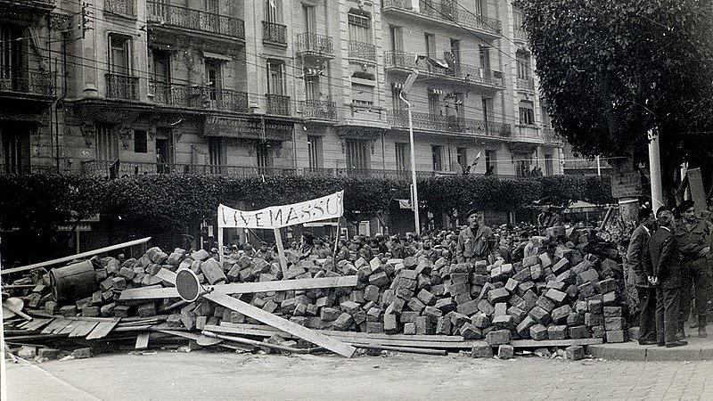 L'image montre une scène historique avec un amas de briques et de débris, suggérant une barricade ou un barrissement. Au premier plan, on peut voir des personnes rassemblées, probablement des manifestants ou des membres d'une foule, observant la scène. Un panneau est visible, ce qui pourrait indiquer le message ou le motif de la manifestation. À l'arrière-plan, des bâtiments avec des balcons sont présents, typiques d'une architecture urbaine. L'atmosphère semble tendue, reflétant un moment de contestation ou de protestation.